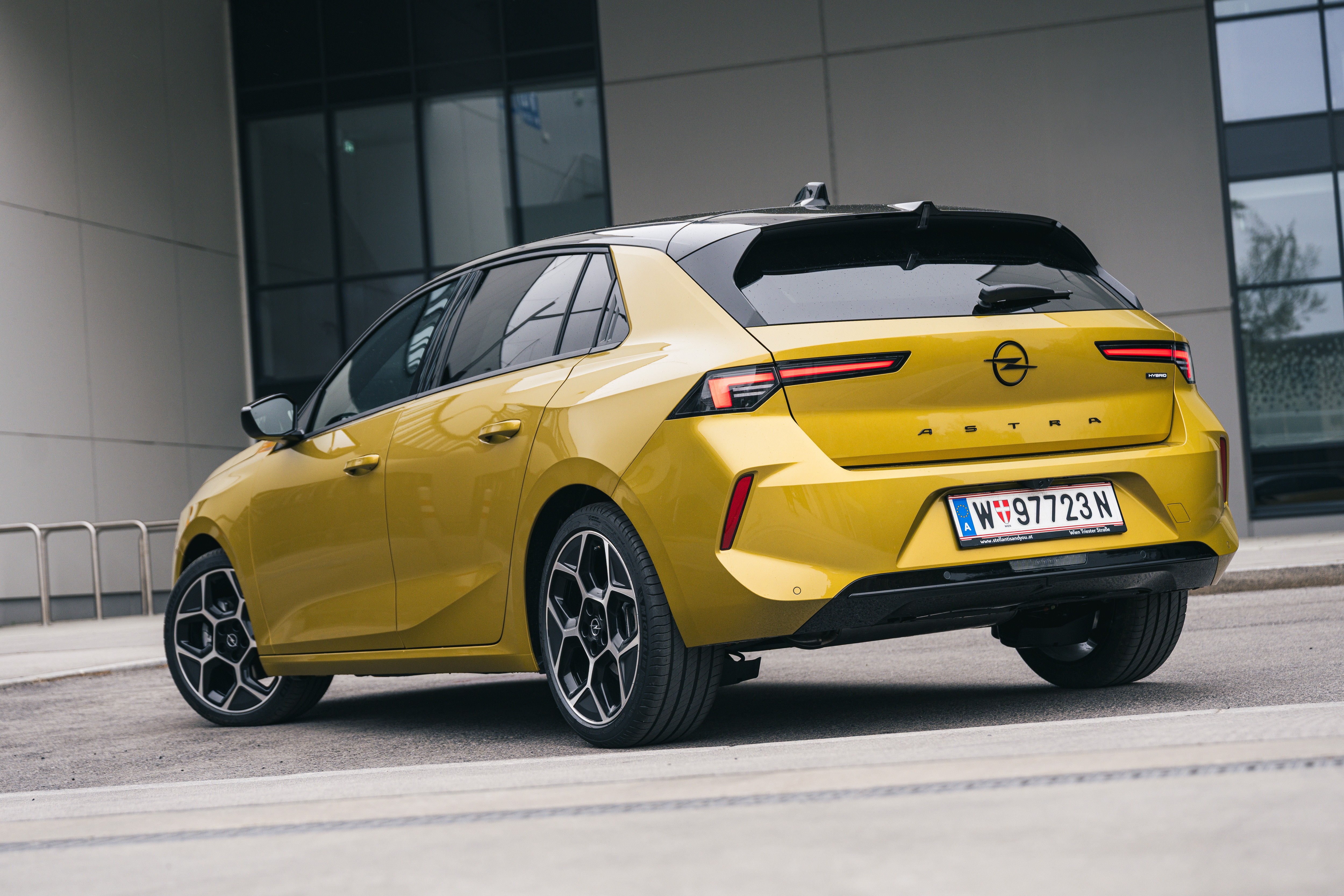 Carro Opel Astra hatchback amarelo com teto preto, visto de traseira e lateral, estacionado em rua de asfalto cinza com pr&eacute;dio moderno ao fundo