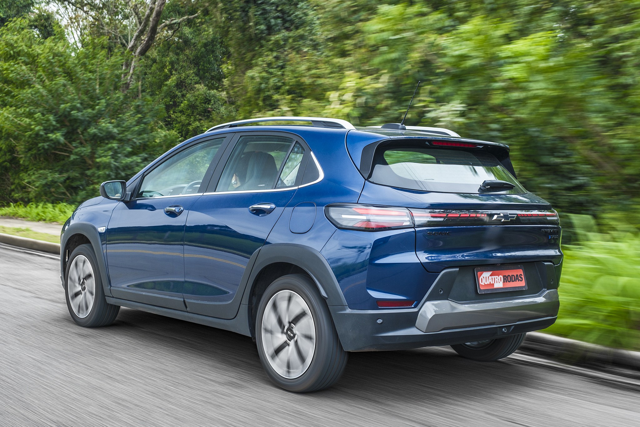Carro azul escuro, modelo SUV compacto, visto de traseira e lateral, em movimento numa estrada com &aacute;rvores verdes ao fundo. As rodas est&atilde;o borradas, indicando velocidade.