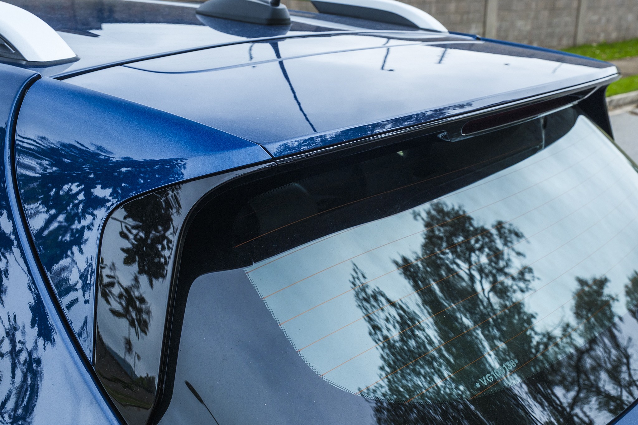 Parte traseira de um carro azul met&aacute;lico, com o vidro traseiro refletindo &aacute;rvores e o c&eacute;u. Um aerof&oacute;lio preto se estende sobre o vidro, e barras de teto brancas s&atilde;o vis&iacute;veis no topo