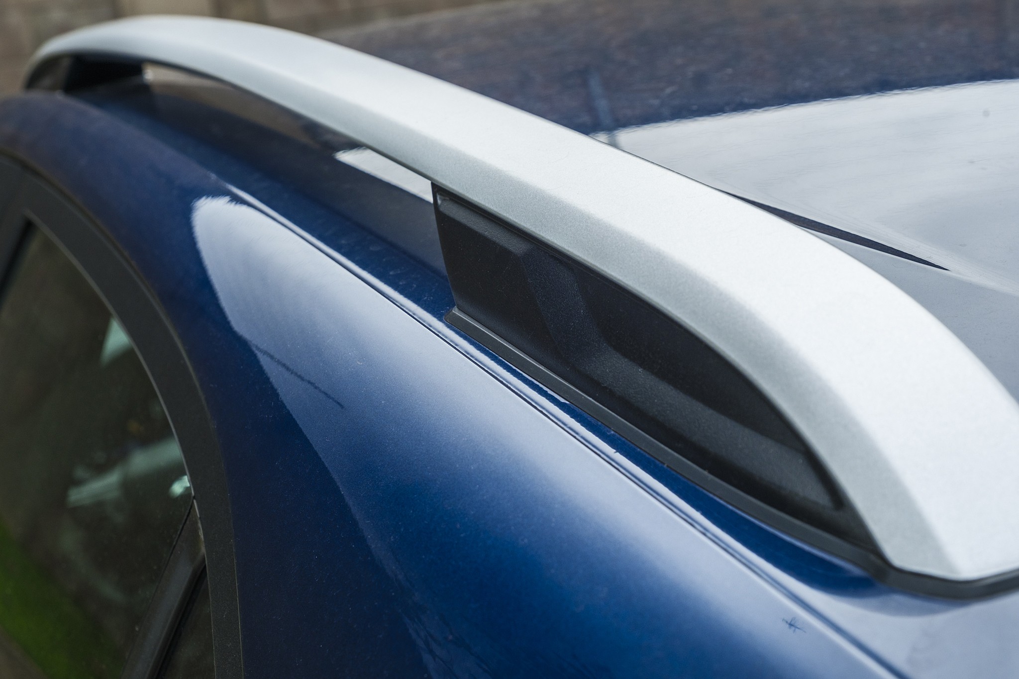 Detalhe do rack de teto prateado de um carro azul escuro, com reflexos de luz na pintura e vidro da janela lateral