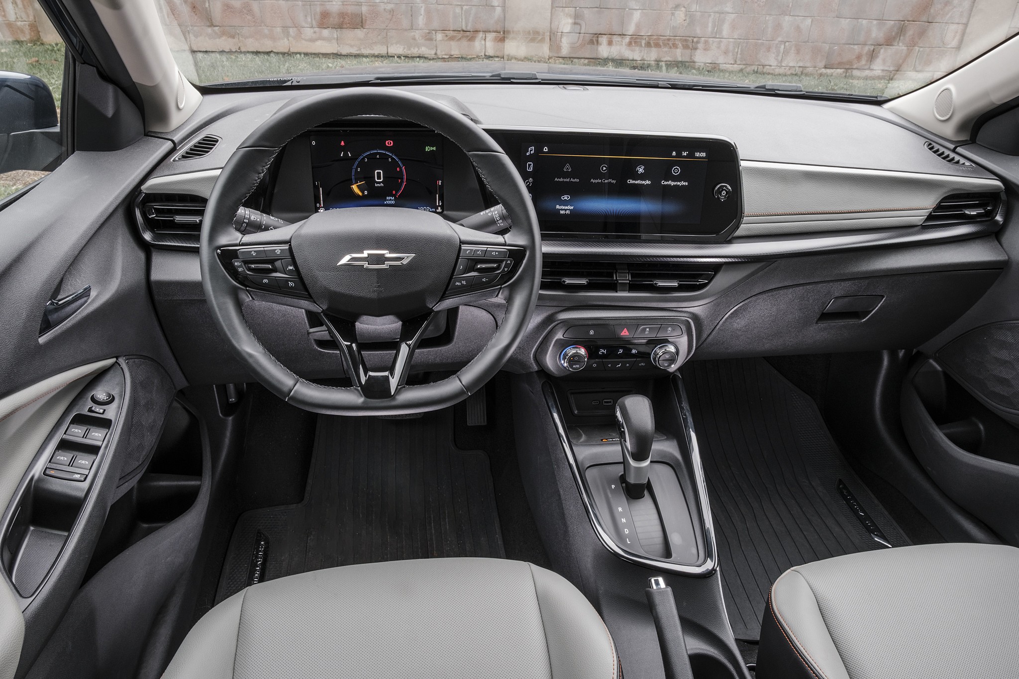 Interior de um carro moderno, com volante multifuncional preto e logo Chevrolet ao centro. Painel digital exibe veloc&iacute;metro e informa&ccedil;&otilde;es do ve&iacute;culo. &Agrave; direita, tela multim&iacute;dia sens&iacute;vel ao toque. C&acirc;mbio autom&aacute;tico e controles de ar-condicionado est&atilde;o no console central. Bancos claros e detalhes em cinza no painel.