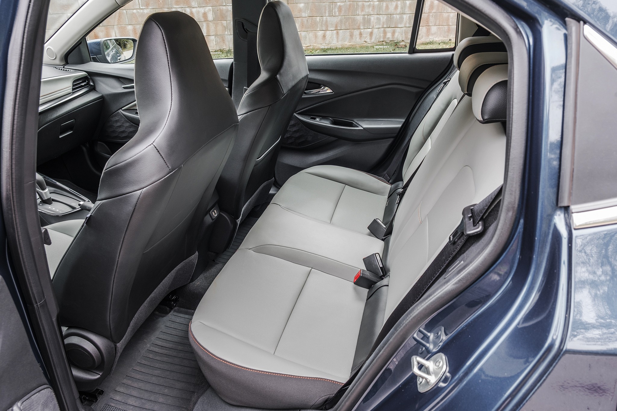 Interior de um carro azul-escuro, mostrando o banco traseiro cinza-claro com detalhes em cinza-escuro e costura vermelha. Os cintos de seguran&ccedil;a est&atilde;o vis&iacute;veis, e os bancos dianteiros pretos s&atilde;o vistos parcialmente. O assoalho tem tapetes de borracha pretos. A janela traseira revela uma parede de tijolos ao fundo