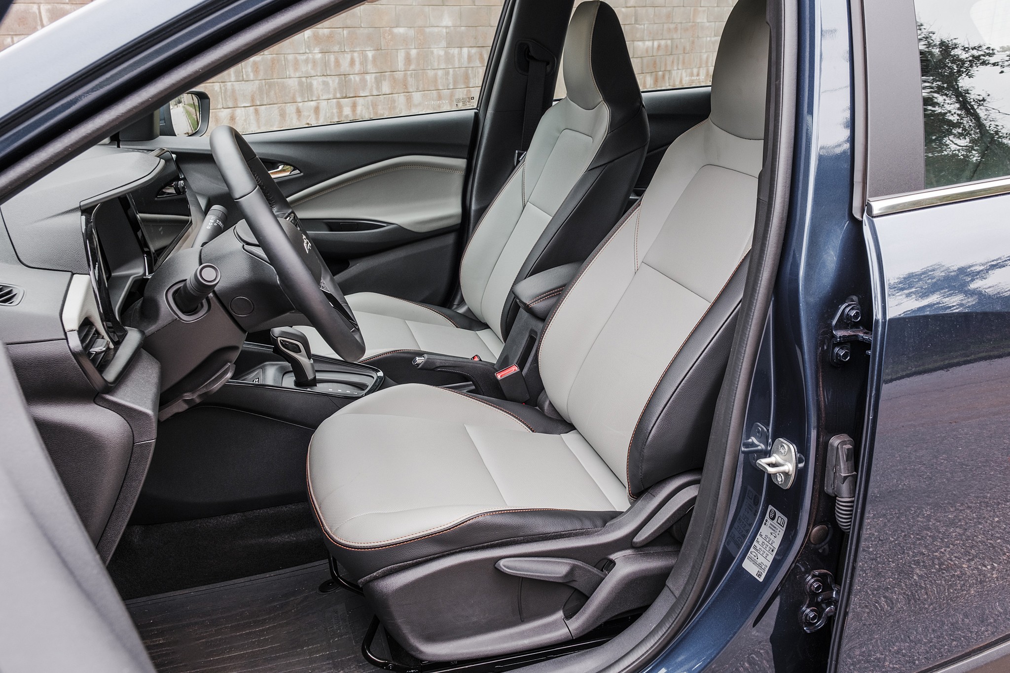 Interior de um carro com bancos dianteiros em couro cinza claro e detalhes pretos, volante escuro e painel com tela central. A porta do motorista est&aacute; aberta, revelando a lateral do ve&iacute;culo em azul escuro. Ao fundo, uma parede de tijolos claros.