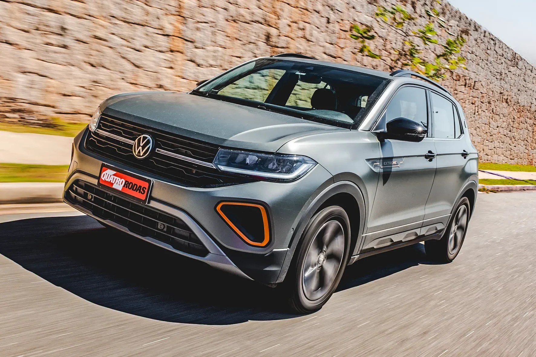 SUV Volkswagen T-Cross cinza-claro em movimento, visto de frente e lateralmente, com detalhes laranja no para-choque. O carro está em uma estrada asfaltada, com um muro de pedras ao fundo e vegetação borrada, indicando velocidade
