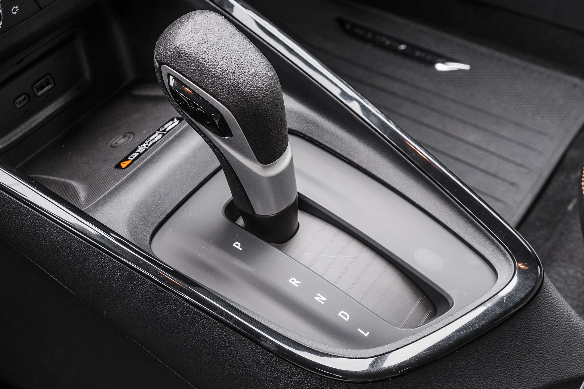 C&acirc;mbio autom&aacute;tico de um carro, com a alavanca em posi&ccedil;&atilde;o neutra, mostrando as letras P, R, N, D e L. Ao lado, h&aacute; um compartimento com duas entradas USB e um &iacute;cone de carregamento sem fio