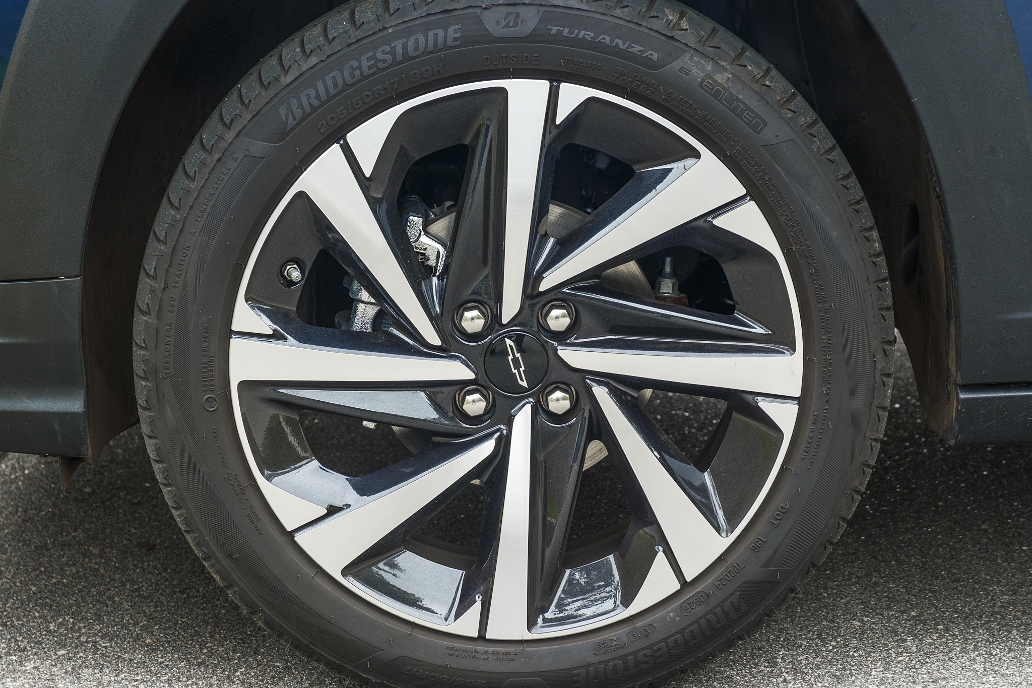 Roda de carro com aro de liga leve preto e prata, pneu Bridgestone Turanza com banda de rodagem vis&iacute;vel, e o emblema da Chevrolet no centro. O para-lama escuro do ve&iacute;culo aparece na parte superior, e o asfalto cinza na parte inferior