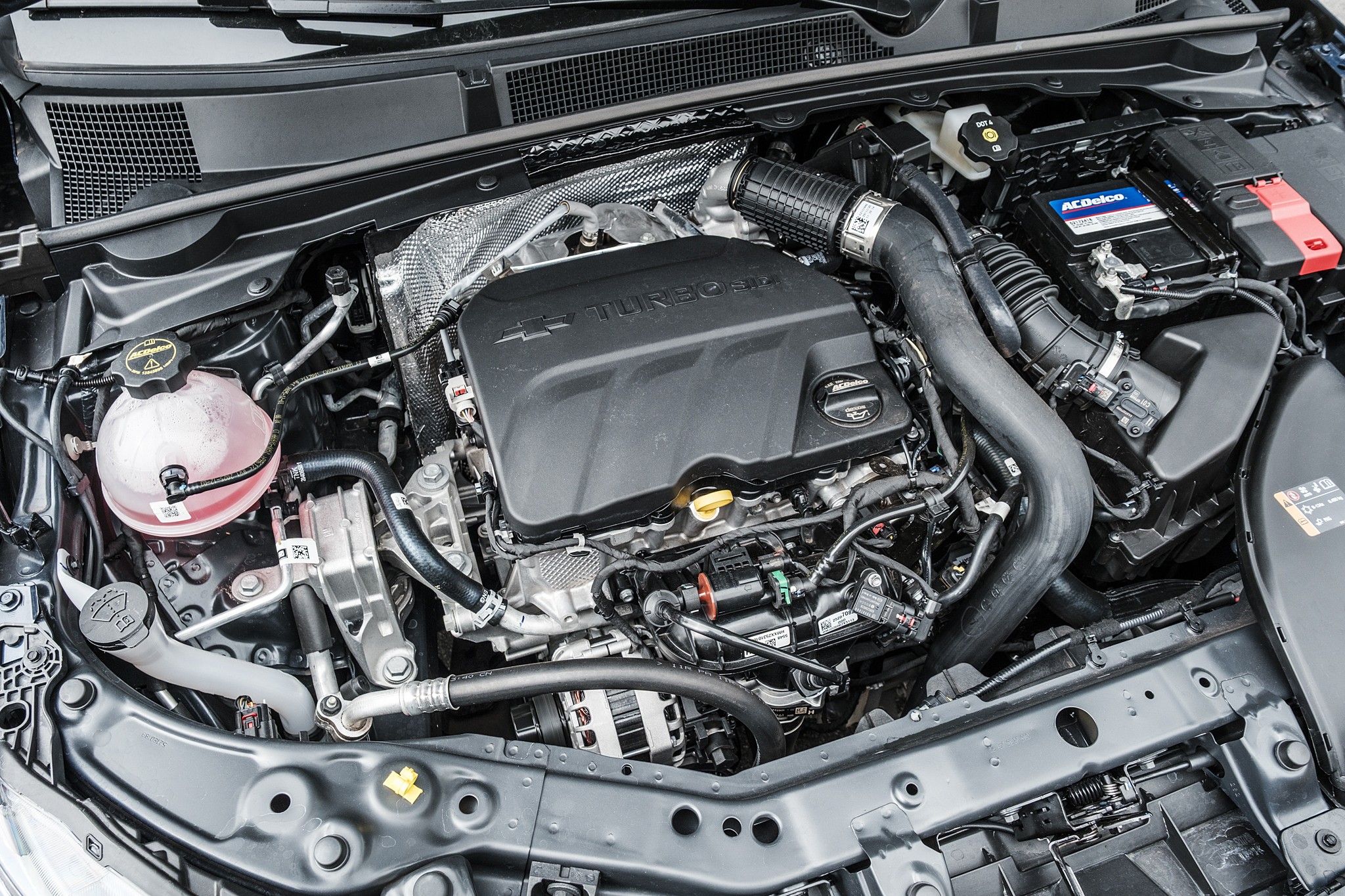 Motor de carro moderno, com a tampa do motor preta em destaque, mangueiras, fios e reservat&oacute;rios vis&iacute;veis, incluindo um reservat&oacute;rio de fluido rosa transl&uacute;cido e uma bateria ACDelco azul