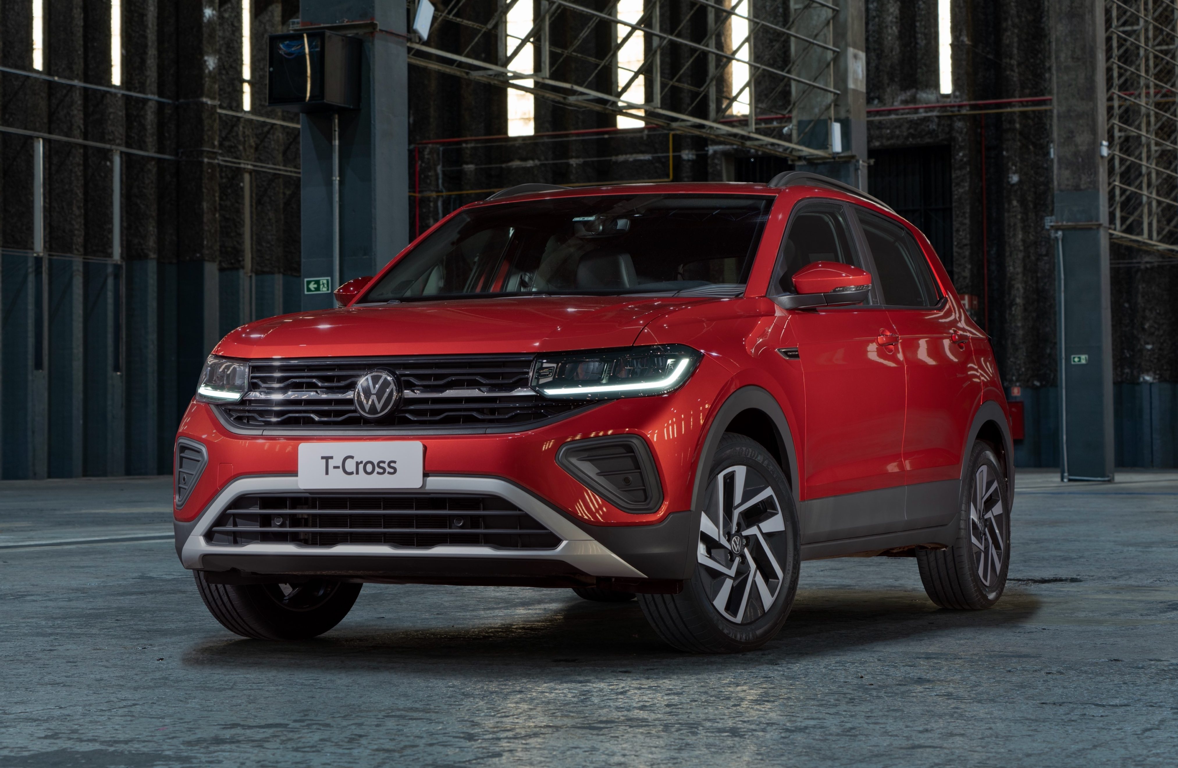 SUV Volkswagen T-Cross vermelho vibrante, com far&oacute;is e luzes diurnas acesas, estacionado em um galp&atilde;o industrial escuro com paredes texturizadas e ilumina&ccedil;&atilde;o vertical