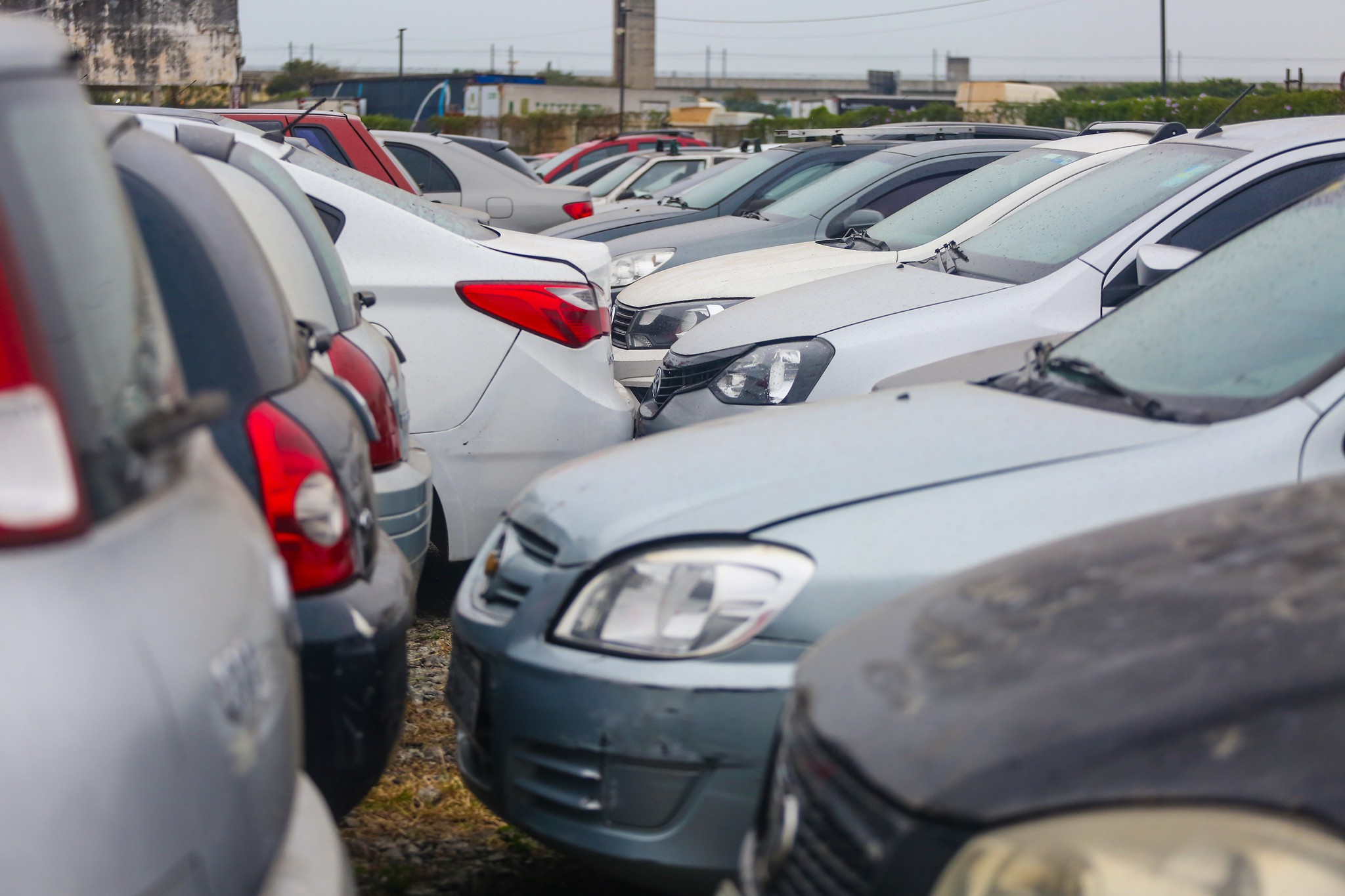 Fileira de carros de diferentes modelos e cores, como branco, cinza e azul, estacionados lado a lado em um p&aacute;tio de terra batida. Os ve&iacute;culos parecem usados, alguns com sujeira e poeira, e est&atilde;o bem pr&oacute;ximos uns dos outros. Ao fundo, estruturas de concreto e vegeta&ccedil;&atilde;o emba&ccedil;adas sob um c&eacute;u nublado