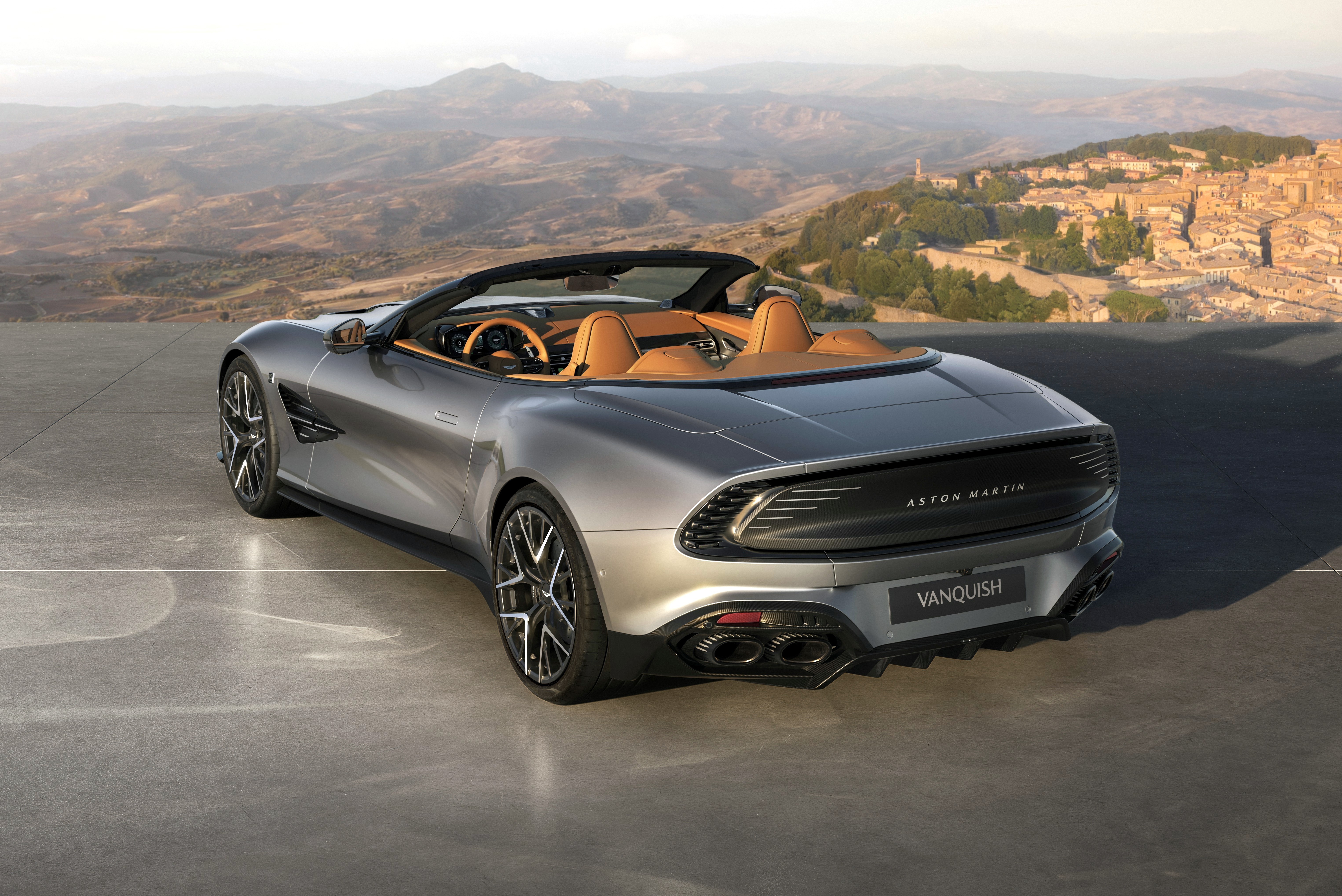 Carro esportivo Aston Martin Vanquish cinza-prateado conversível, com interior de couro marrom, estacionado em um mirante com vista para montanhas e uma cidade antiga ao fundo