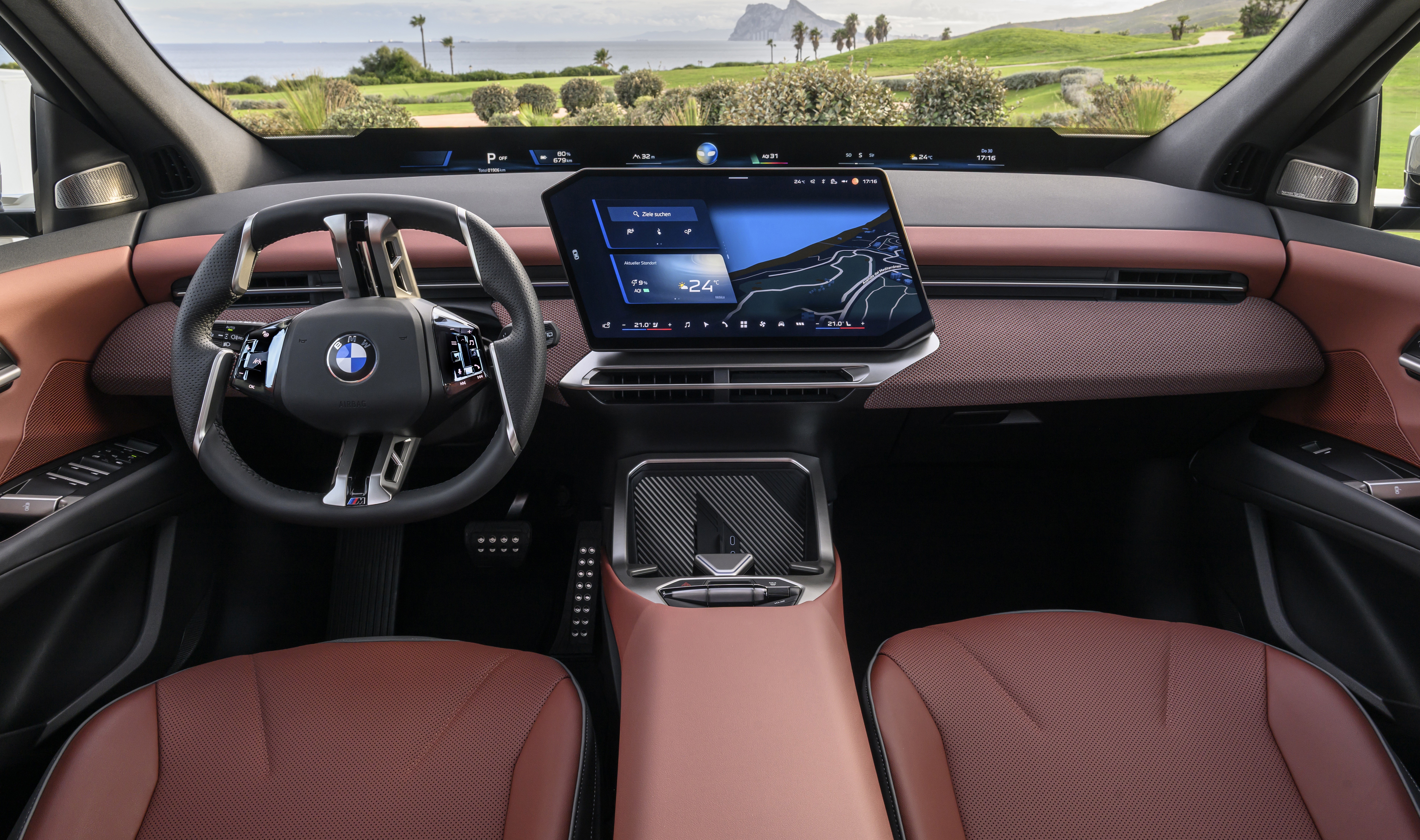 Interior de um carro moderno com volante BMW, painel marrom e tela central exibindo mapa e temperatura. Bancos de couro marrom e paisagem verde com mar ao fundo vis&iacute;vel pelo para-brisa