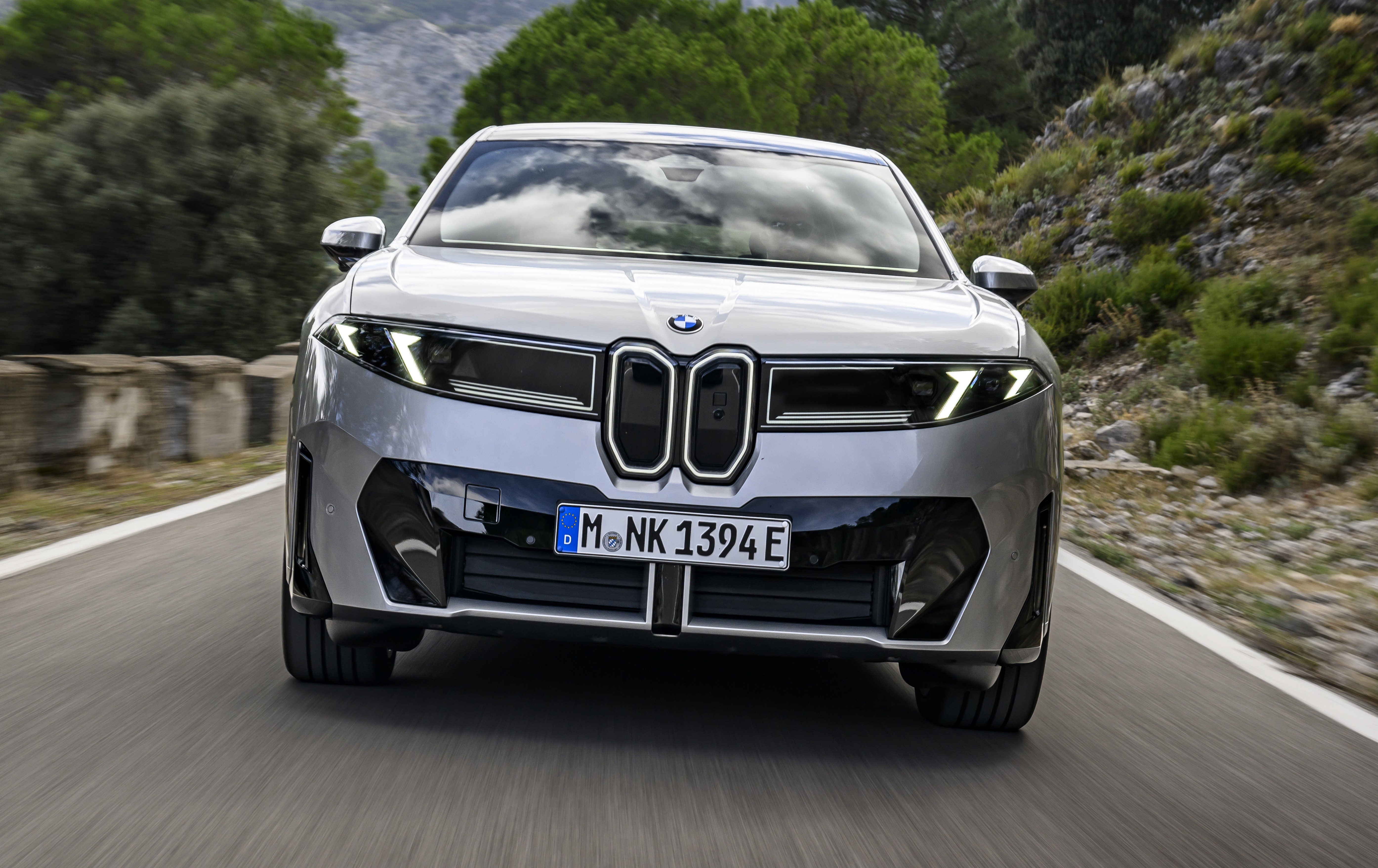 Carro BMW prateado em movimento, visto de frente, com far&oacute;is acesos e grade dupla em destaque, em uma estrada sinuosa com vegeta&ccedil;&atilde;o e montanhas ao fundo
