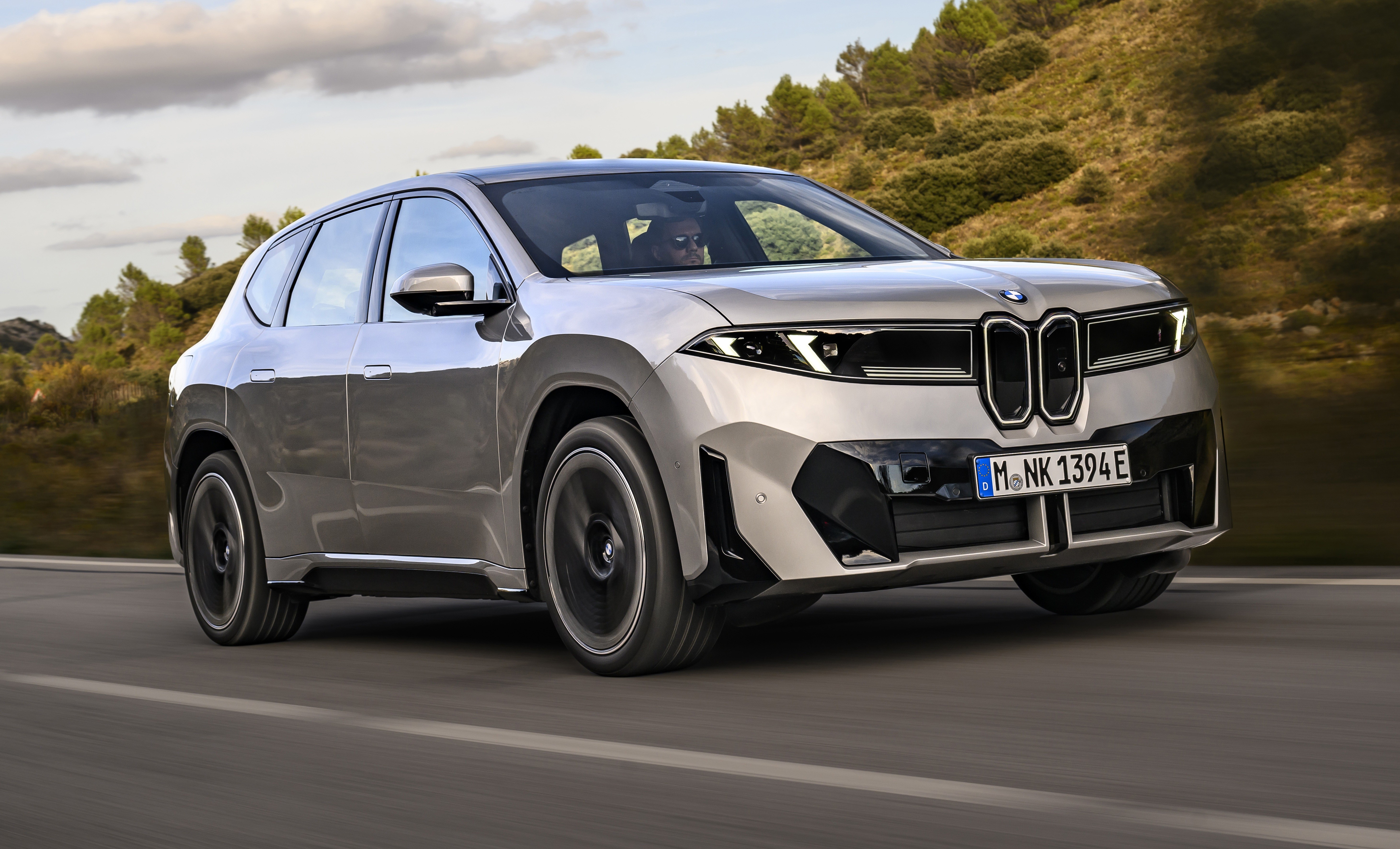 SUV BMW iX prateado em movimento r&aacute;pido numa estrada, com montanhas e c&eacute;u nublado ao fundo
