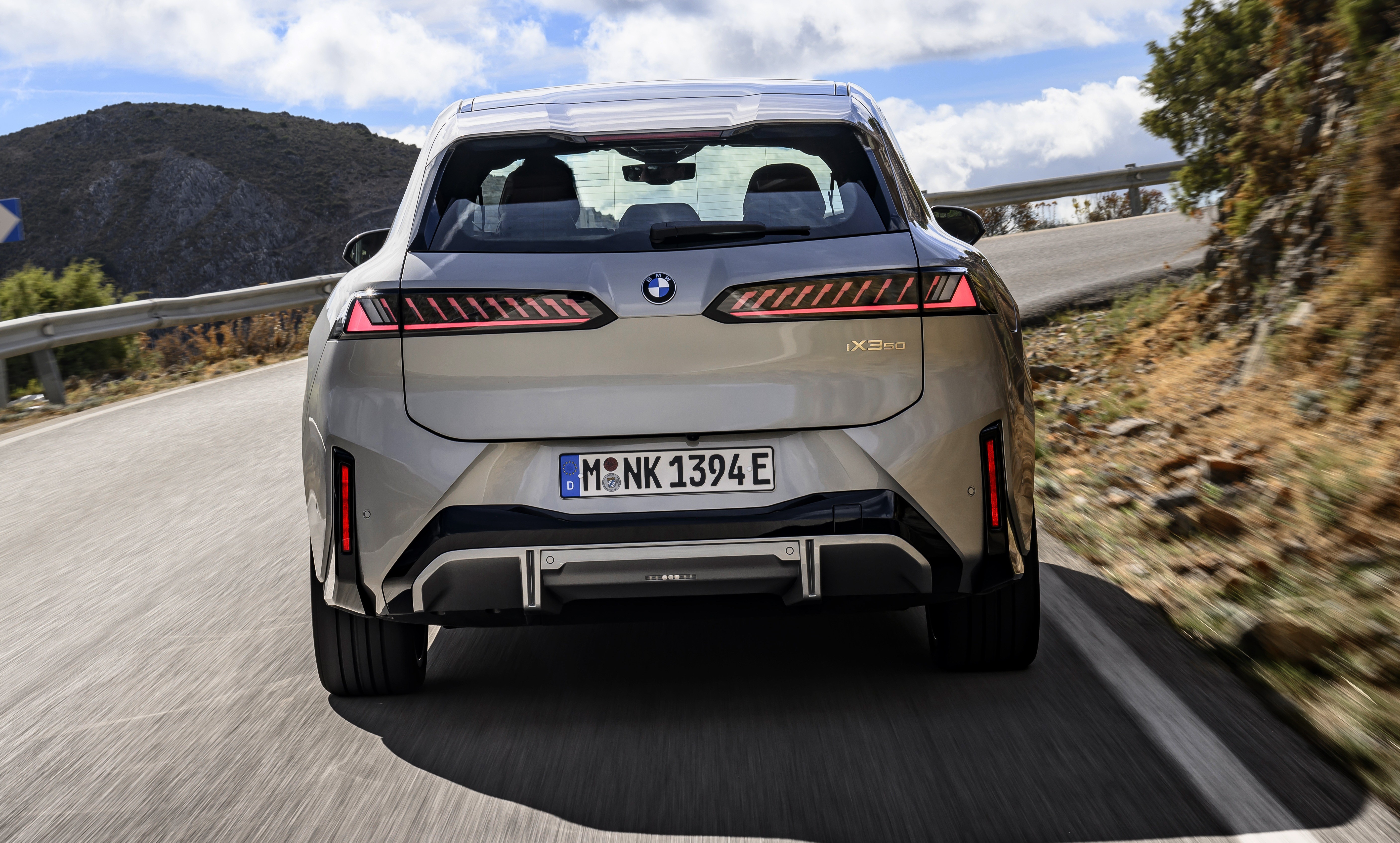 Traseira de um carro BMW iX3 el&eacute;trico cinza claro em uma estrada sinuosa, com montanhas e c&eacute;u azul com nuvens ao fundo