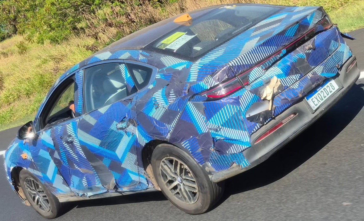 Carro camuflado com adesivos azuis e pretos em padr&atilde;o geom&eacute;trico, visto de traseira e lateral, em uma estrada com vegeta&ccedil;&atilde;o ao fundo. A placa traseira &eacute; EXQ2D28