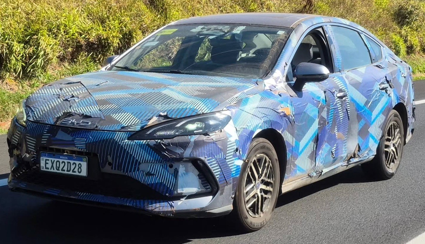 Carro de teste camuflado em tons de azul e preto, com padr&atilde;o geom&eacute;trico, trafegando em uma estrada. O ve&iacute;culo tem placa brasileira EXQ2D28 e far&oacute;is acesos, com vegeta&ccedil;&atilde;o ao fundo
