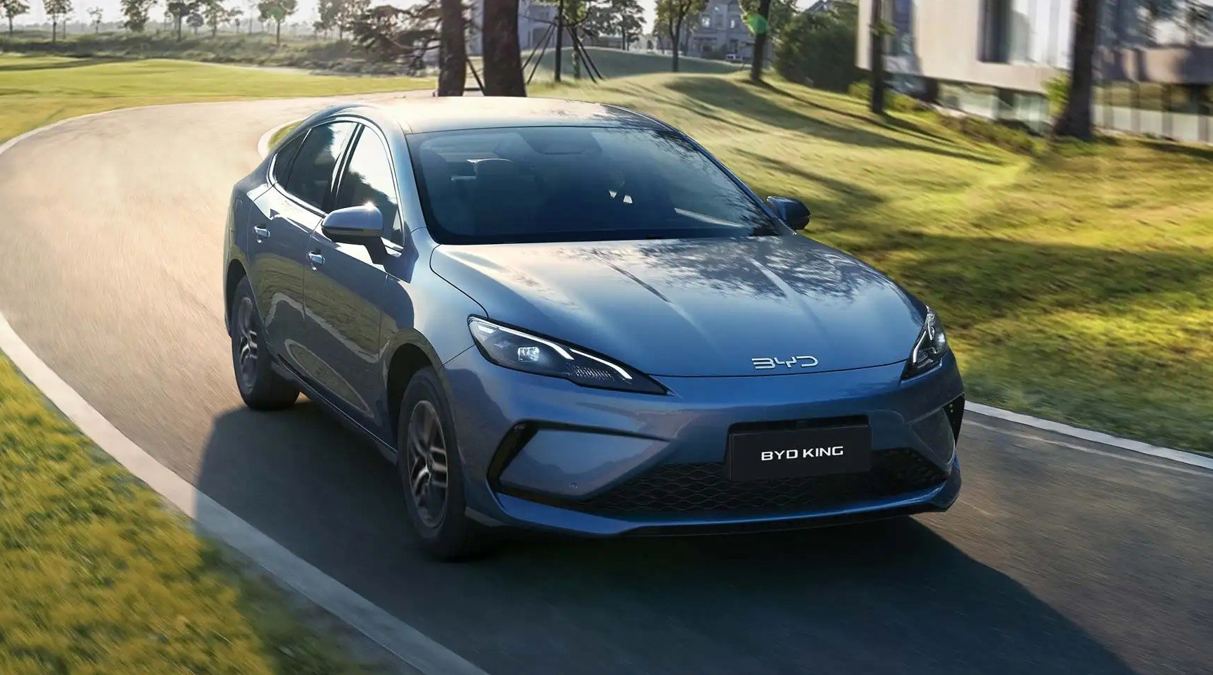 Sedan BYD King azul met&aacute;lico em uma curva suave de estrada asfaltada, com grama verde e &aacute;rvores ao fundo, sob luz solar