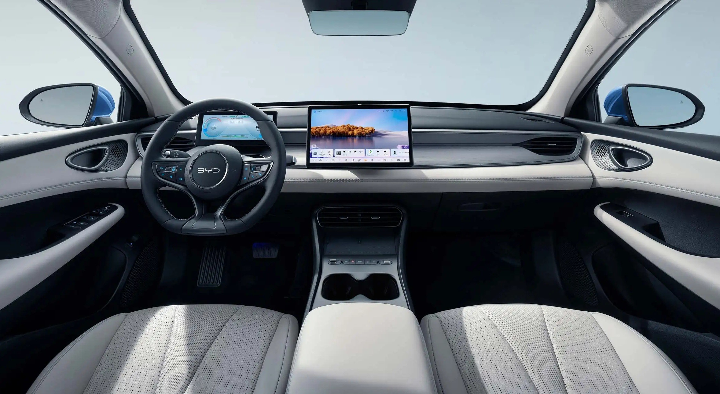 Interior de um carro moderno com bancos claros, volante multifuncional escuro e uma grande tela central exibindo uma paisagem com &aacute;rvores e &aacute;gua. O painel &eacute; cinza claro e h&aacute; um console central com porta-copos.
