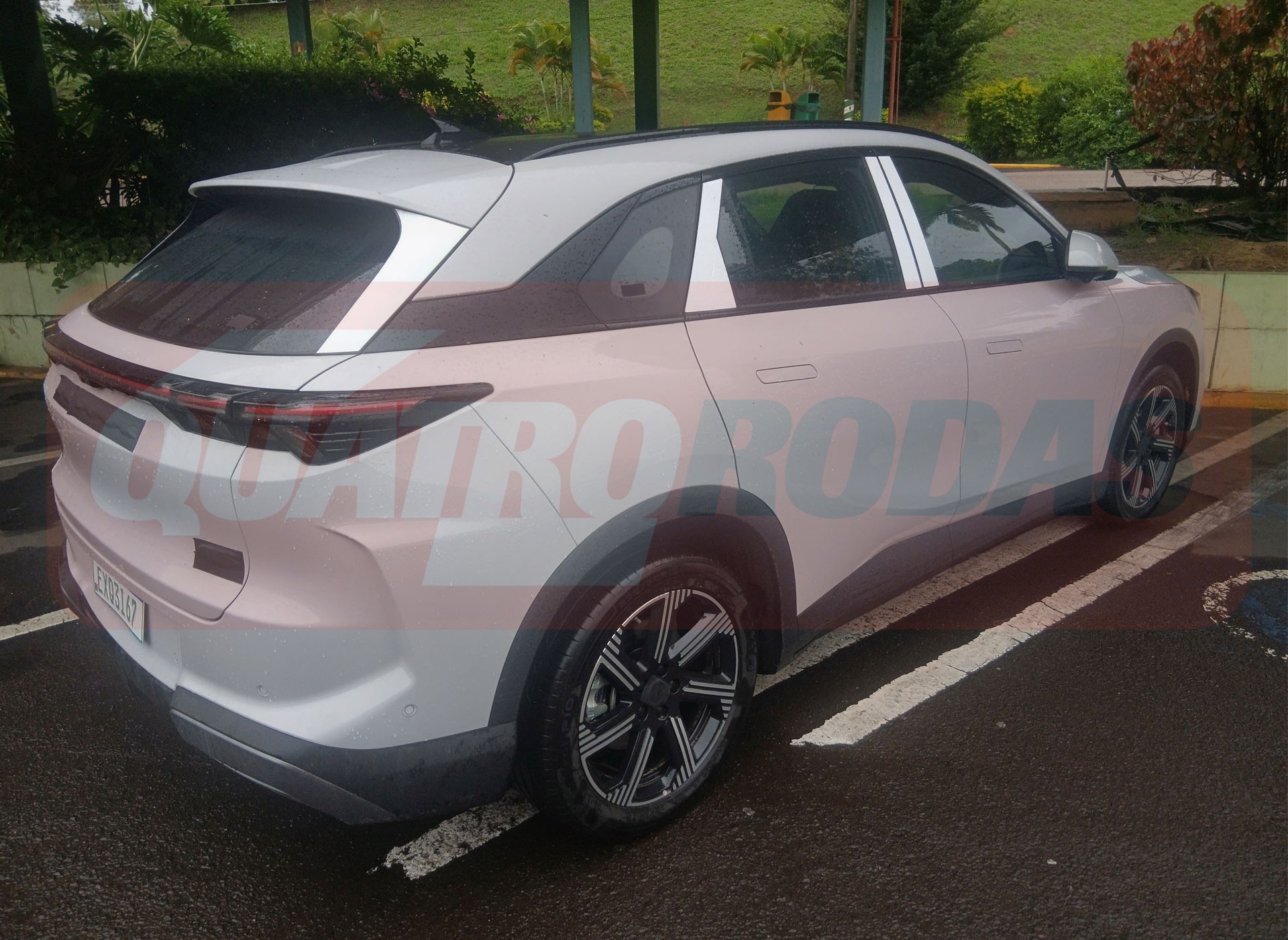 SUV branco estacionado em vaga de rua, visto de tr&aacute;s e lateral direita. Possui lanternas traseiras finas e horizontais, ma&ccedil;anetas embutidas e rodas com design geom&eacute;trico em preto e prata. O para-choque traseiro e as caixas de roda s&atilde;o em pl&aacute;stico escuro. O carro est&aacute; em uma &aacute;rea pavimentada com marca&ccedil;&otilde;es de estacionamento, e h&aacute; vegeta&ccedil;&atilde;o ao fundo