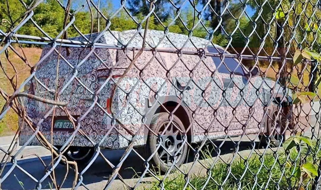 Carro prot&oacute;tipo camuflado com estampa colorida, visto por tr&aacute;s atrav&eacute;s de uma cerca de arame, em uma estrada com vegeta&ccedil;&atilde;o ao fundo