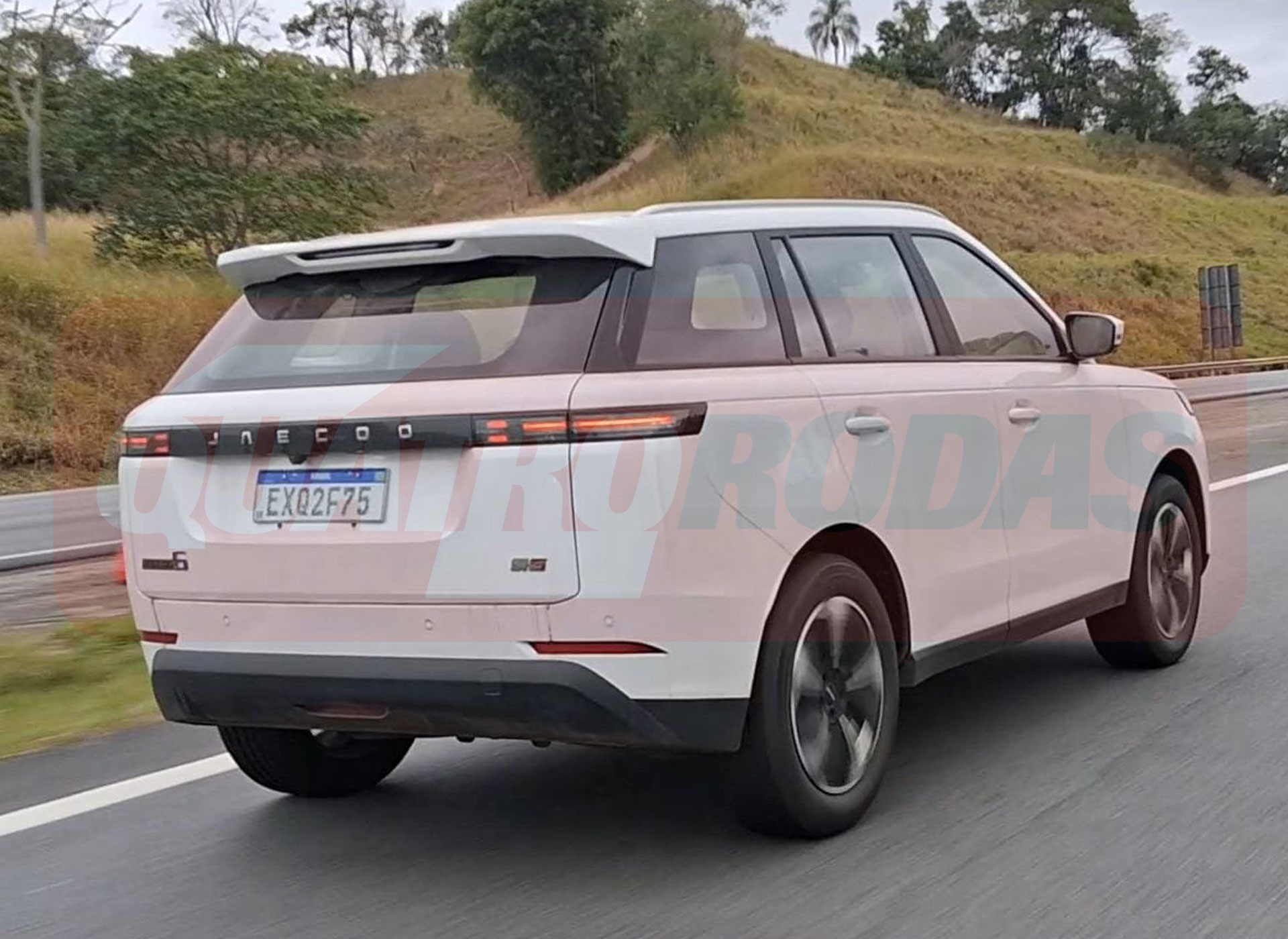SUV branco Jaecoo J6 com placa brasileira EXQ2F75, visto de traseira e lateral, trafegando em rodovia com vegeta&ccedil;&atilde;o ao fundo