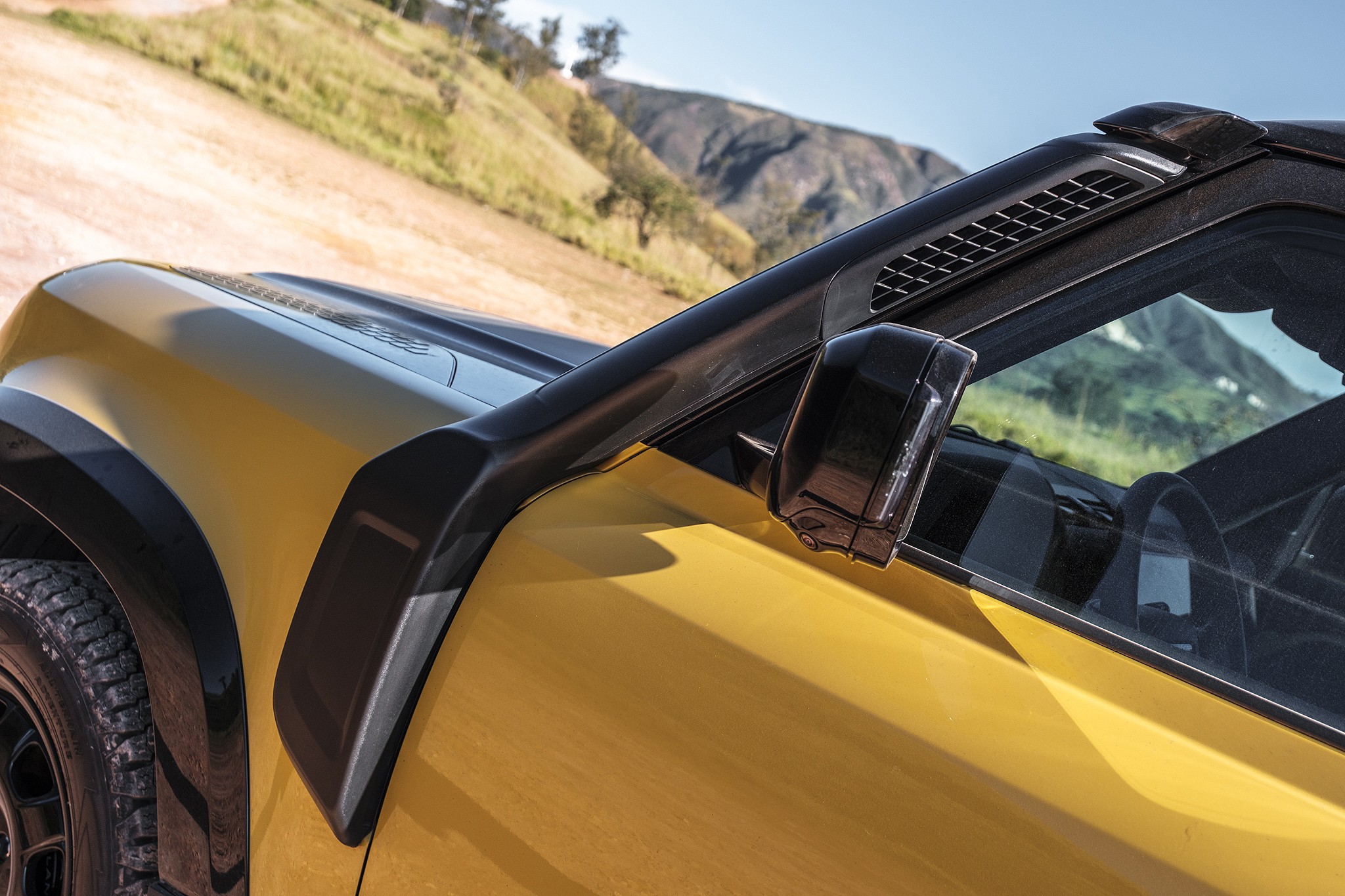 Lateral de um carro amarelo com detalhes pretos, incluindo o para-lama, retrovisor e um snorkel. Ao fundo, uma estrada de terra e montanhas com vegeta&ccedil;&atilde;o rasteira sob c&eacute;u azul