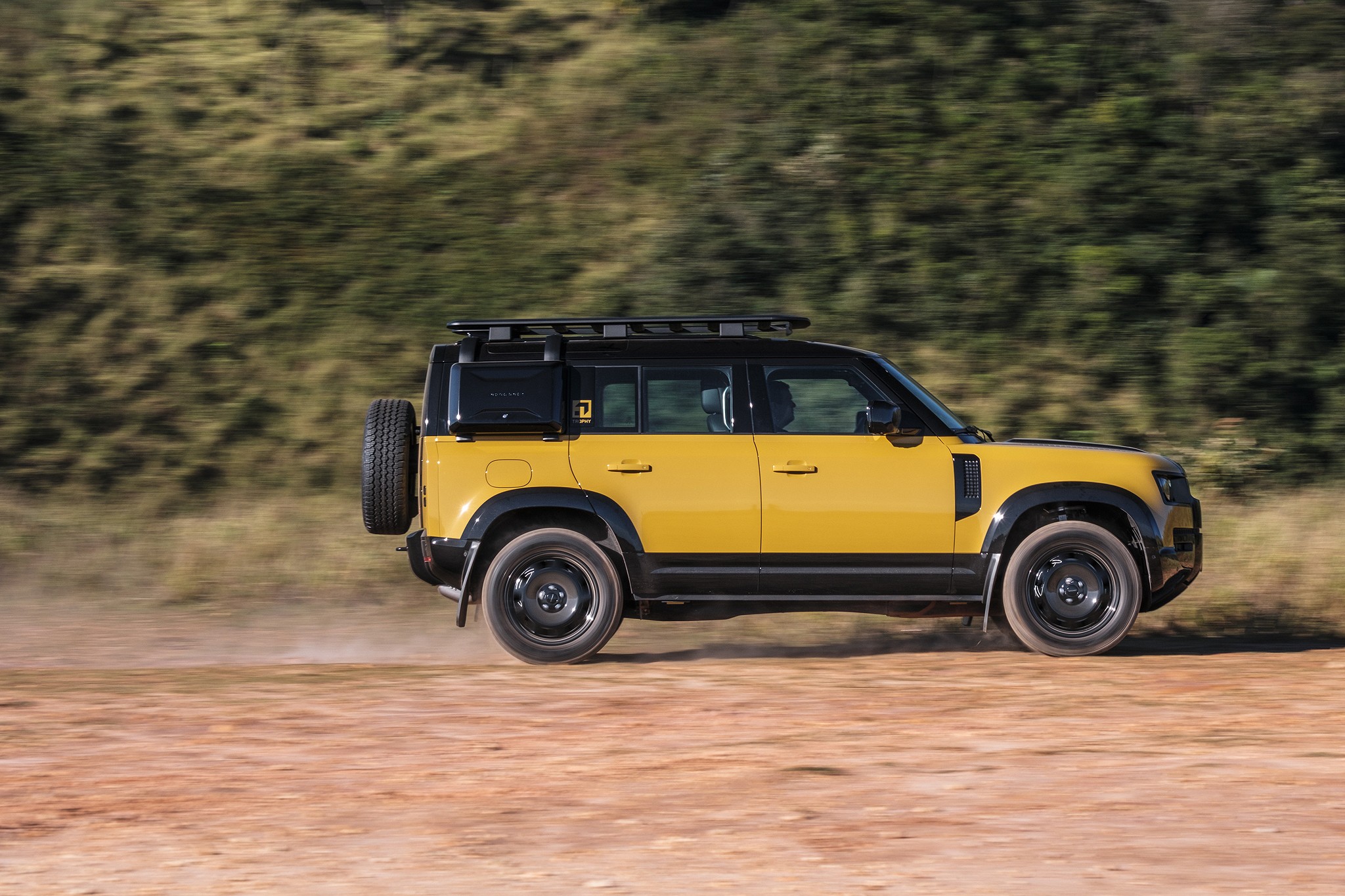 Um Land Rover Defender amarelo e preto em movimento, visto de lado, levantando poeira em uma estrada de terra com vegeta&ccedil;&atilde;o borrada ao fundo