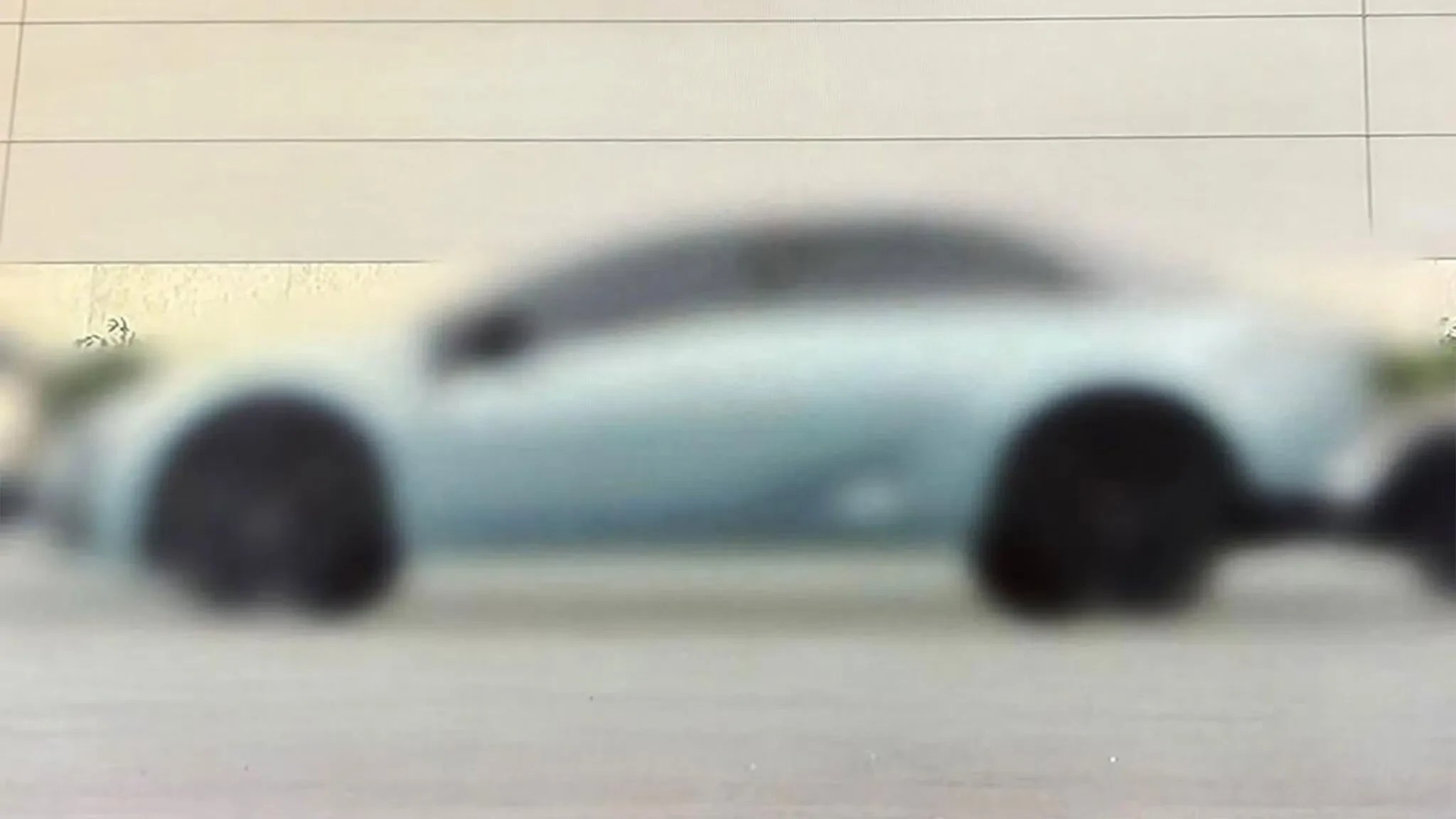 Carro esportivo azul claro, com rodas escuras, estacionado em uma rua ou estacionamento, com um fundo de parede clara e vegetação desfocada
