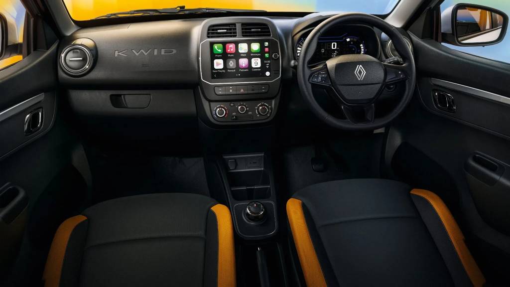 Interior de um carro Renault Kwid, com painel preto e detalhes em laranja nos bancos. O volante tem o logo da Renault, e o painel central exibe uma tela multim&iacute;dia com aplicativos de celular. O nome KWID est&aacute; em relevo no painel do lado do passageiro