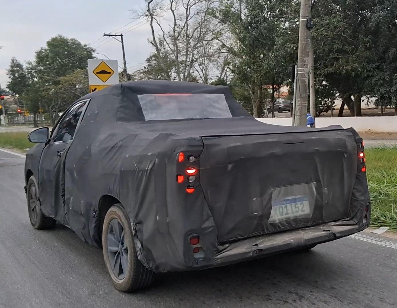 Picape camuflada em testes, vista por tr&aacute;s, com lanternas acesas e placa de teste XQ1I52, rodando em estrada com sinaliza&ccedil;&atilde;o de lombada e &aacute;rvores ao fundo