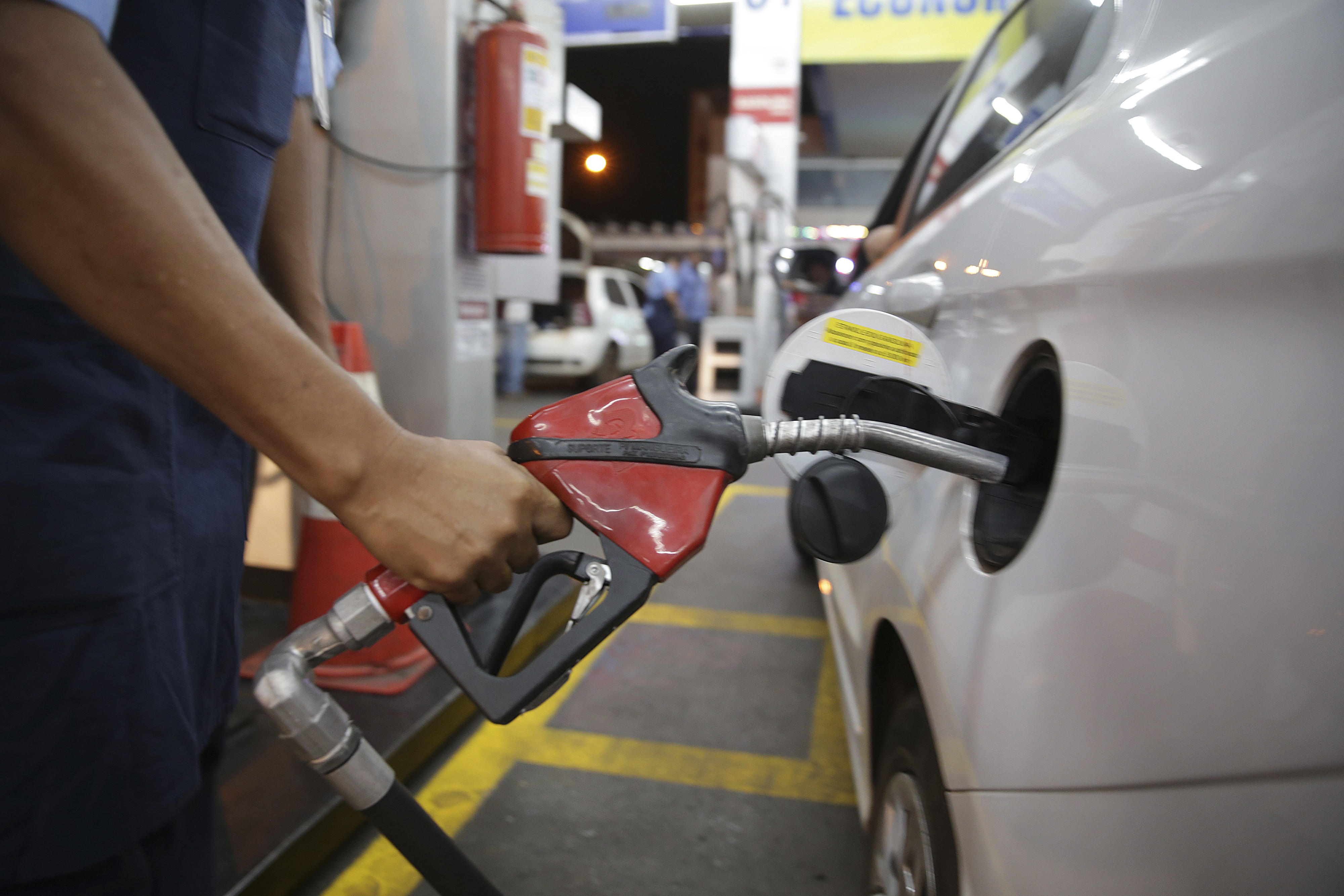 M&atilde;o de um frentista segurando um bico de bomba de combust&iacute;vel vermelho e preto, inserido no tanque de um carro branco em um posto de gasolina &agrave; noite. Ao fundo, um extintor de inc&ecirc;ndio vermelho e outras bombas de combust&iacute;vel