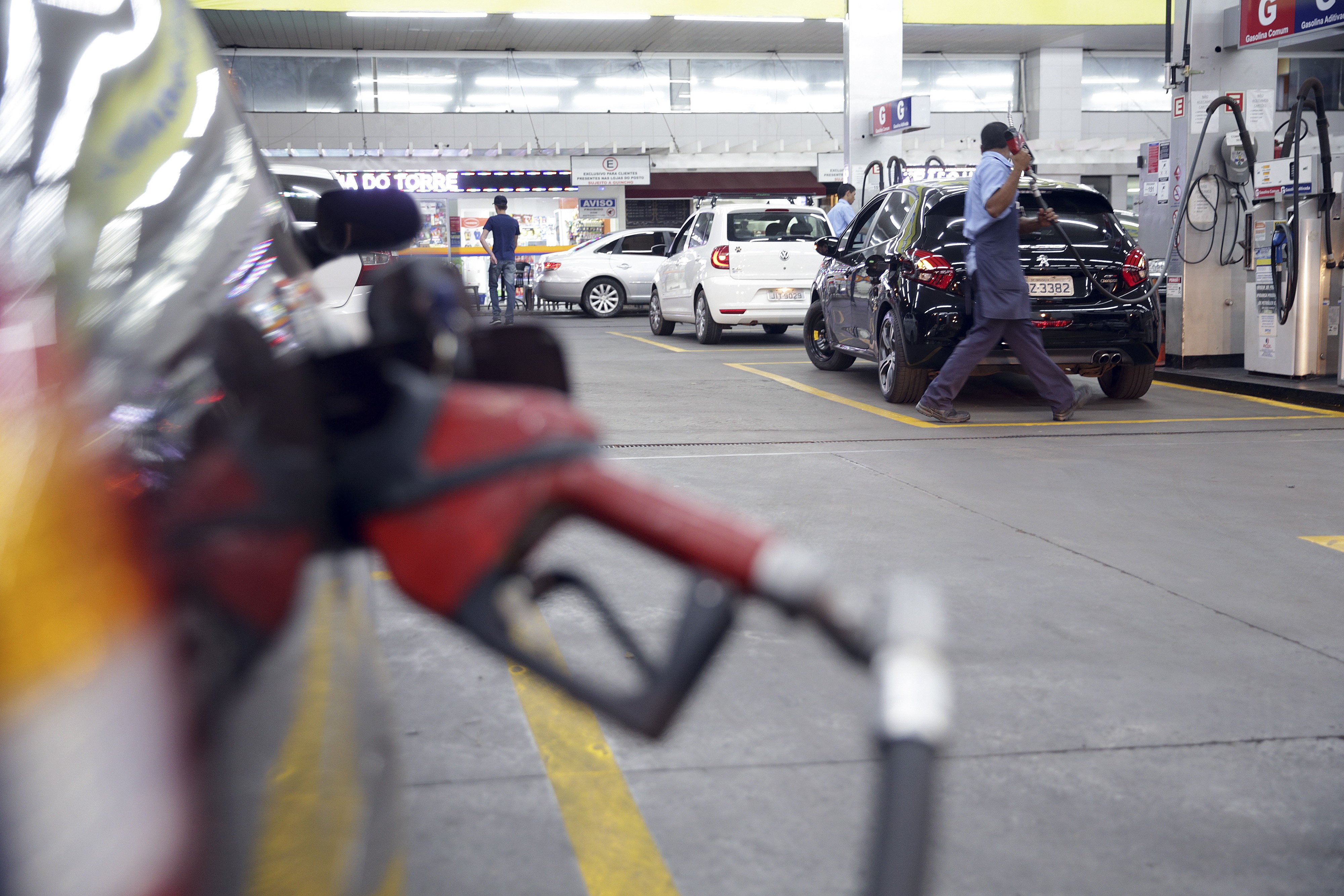 Bico de bomba de combust&iacute;vel vermelho em primeiro plano, desfocado. Ao fundo, um frentista de camisa azul e avental escuro abastece um carro preto em um posto de gasolina movimentado, com outros ve&iacute;culos e pessoas aguardando
