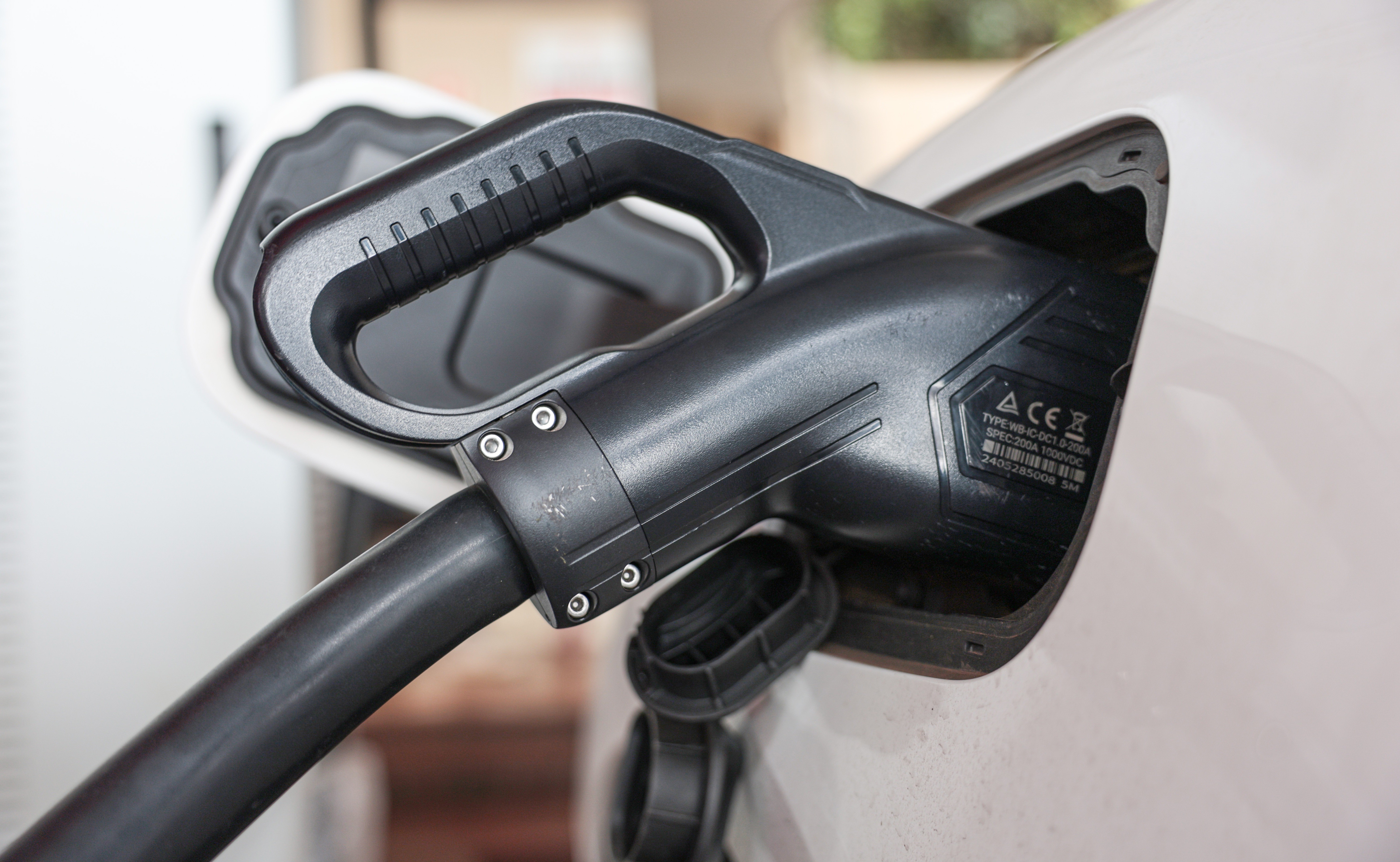Um carregador preto de ve&iacute;culos el&eacute;tricos conectado &agrave; porta de carregamento de um carro branco, com detalhes t&eacute;cnicos vis&iacute;veis no carregador