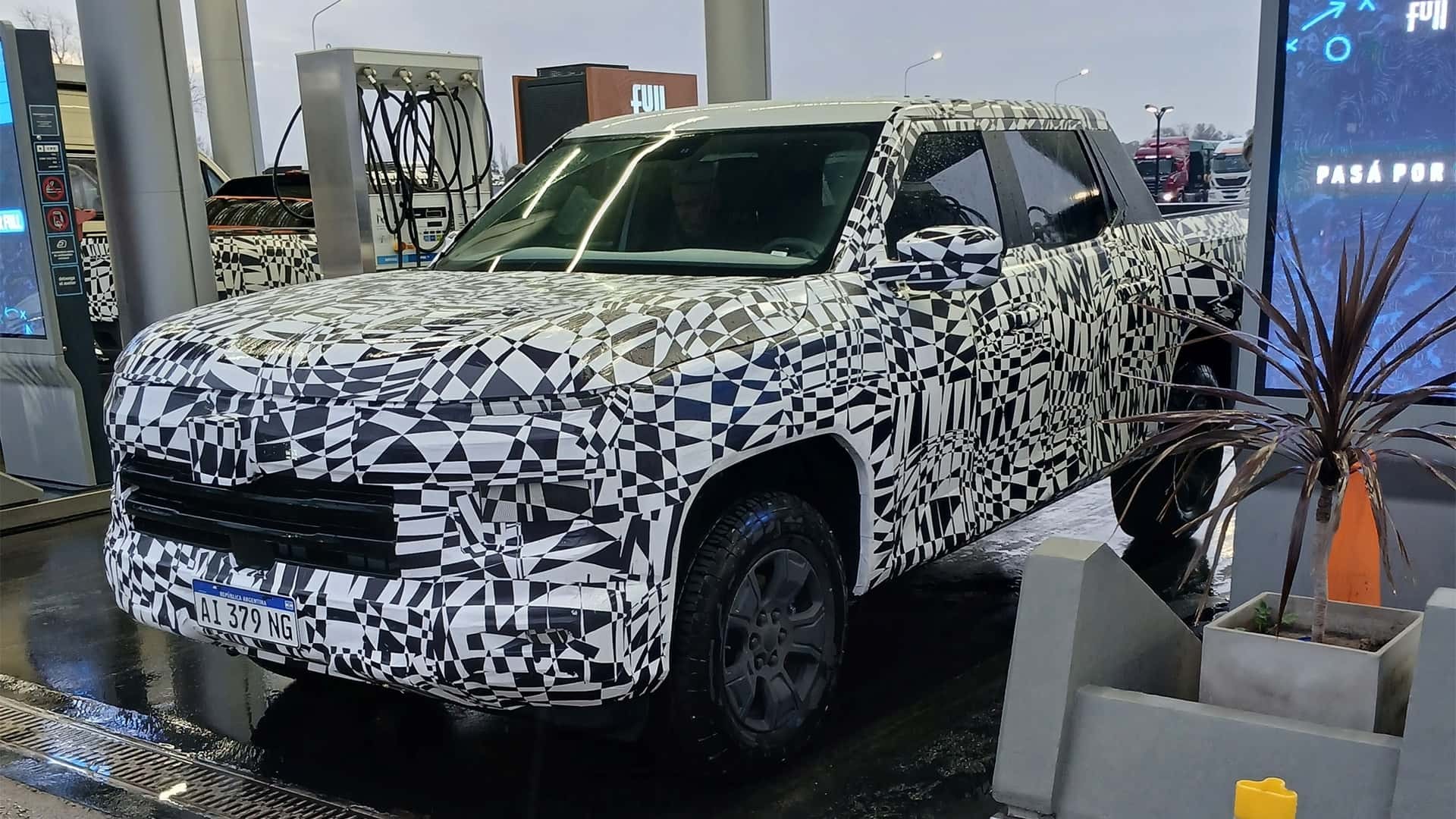 Picape camuflada com adesivos pretos e brancos em padrão geométrico, estacionada em posto de combustível. A placa frontal exibe AL 379 NG. Ao fundo, bombas de gasolina e um painel digital com a frase PASÁ POR. À direita, um vaso com planta de folhas escuras