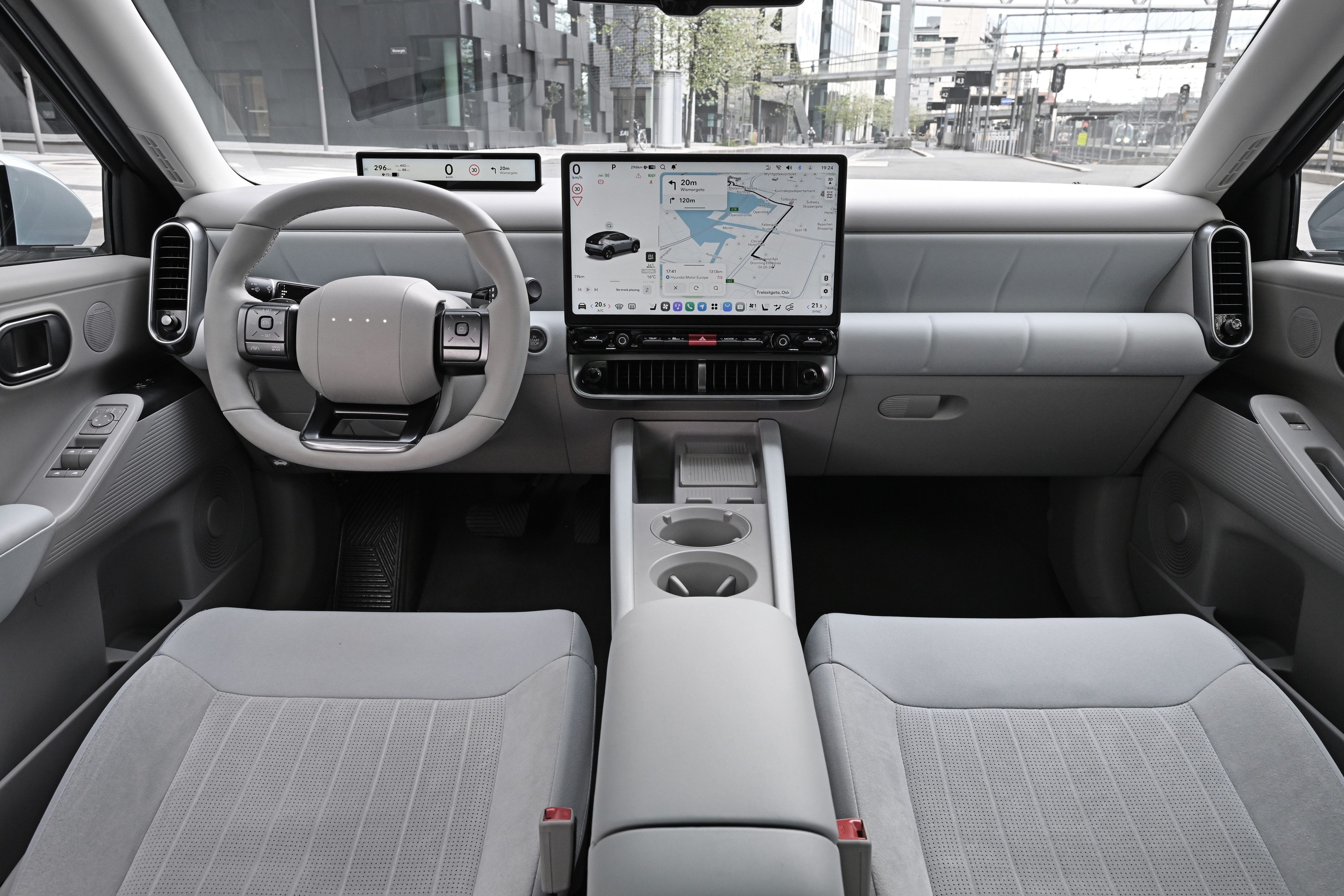 Interior de um carro moderno com acabamento cinza claro. Volante minimalista, painel digital e tela central exibindo um mapa de navega&ccedil;&atilde;o. Bancos dianteiros estofados em tecido cinza com padr&atilde;o quadriculado. Console central com porta-copos.