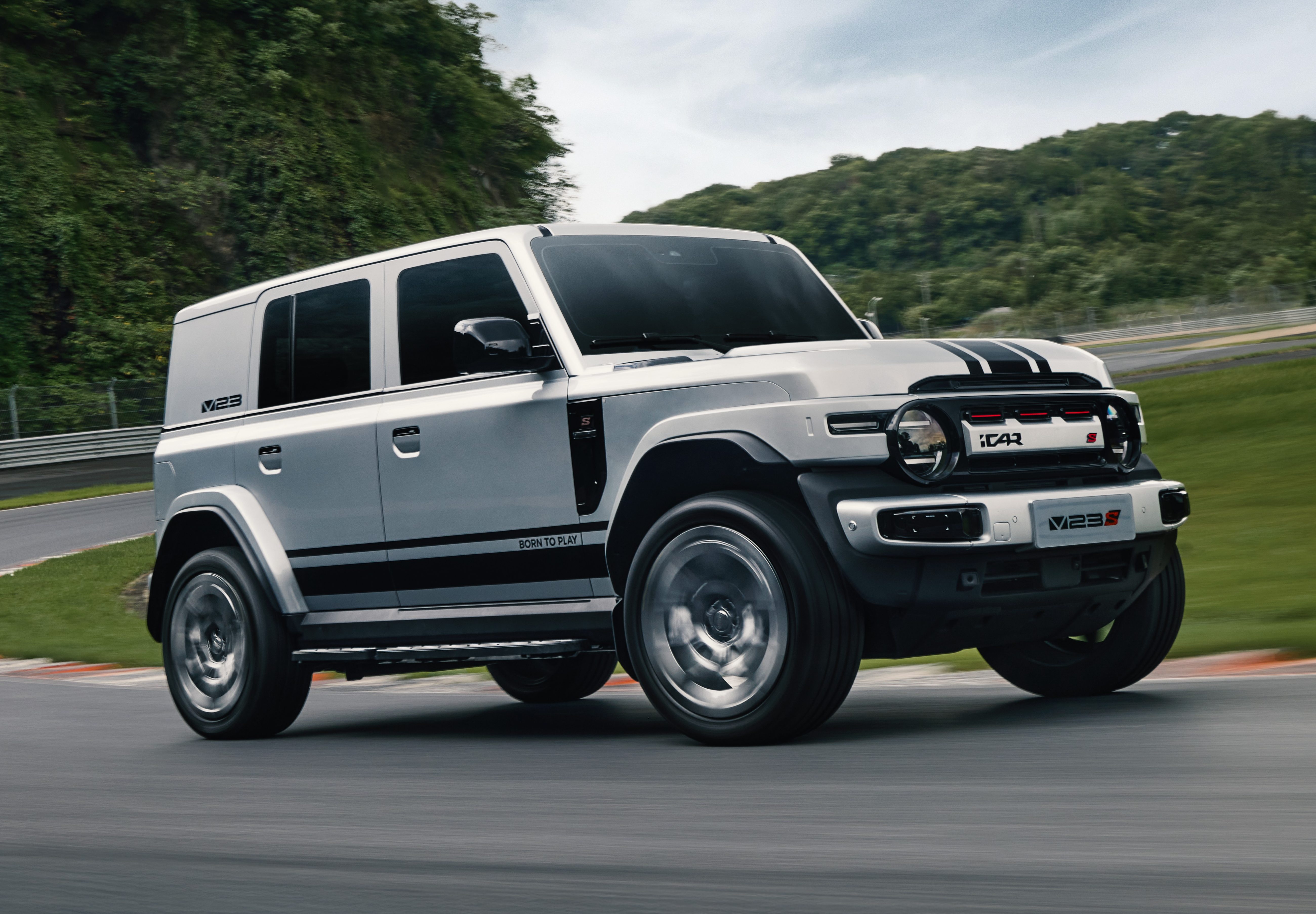 SUV branco com faixas pretas no cap&ocirc; e laterais, em uma curva de pista de corrida, com montanhas verdes ao fundo e c&eacute;u nublado