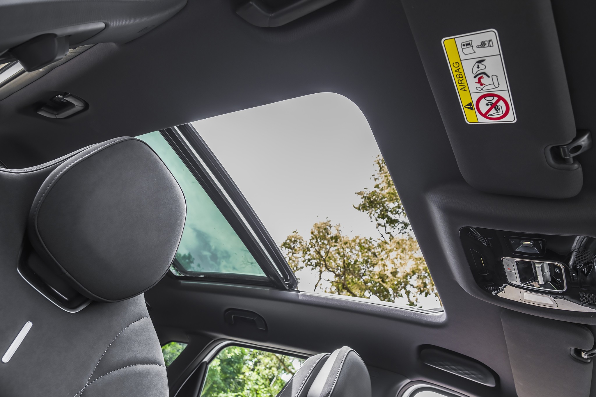 Interior de um carro com teto solar panor&acirc;mico aberto, mostrando o c&eacute;u claro e &aacute;rvores verdes. O encosto de cabe&ccedil;a do banco dianteiro esquerdo, estofado em couro preto com costuras brancas, est&aacute; em primeiro plano. No para-sol direito, um adesivo de aviso de airbag &eacute; vis&iacute;vel