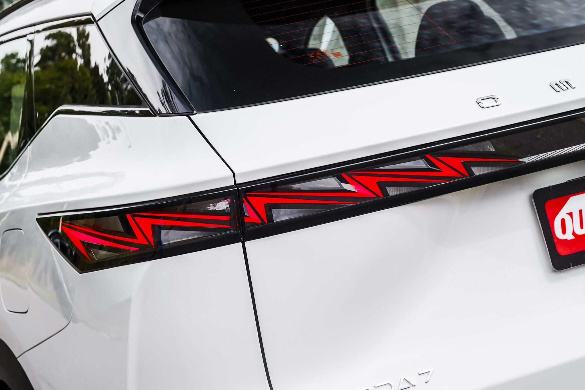 Lanterna traseira de LED vermelha e preta com design em zigue-zague, integrada &agrave; carroceria branca de um carro, com reflexos de &aacute;rvores no vidro traseiro