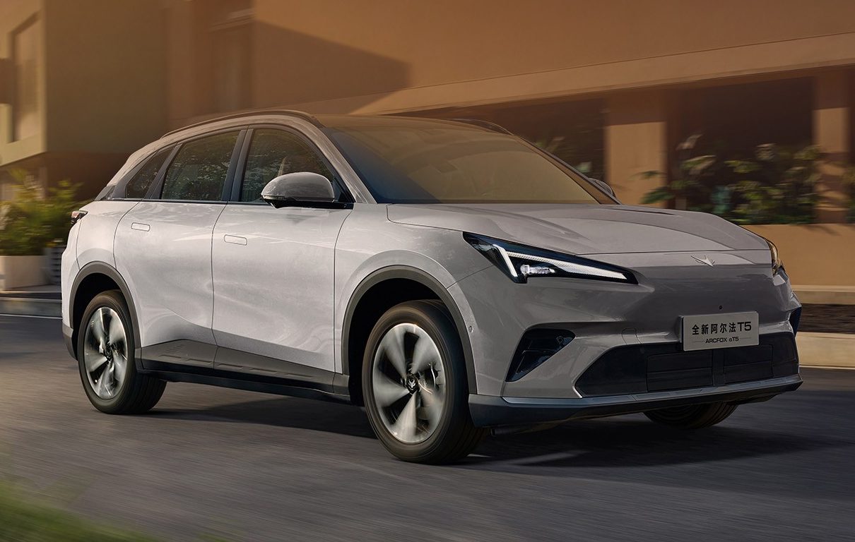 SUV el&eacute;trico cinza claro com detalhes escuros na frente, rodando em uma rua asfaltada com pr&eacute;dios modernos e &aacute;rvores ao fundo, sob luz solar