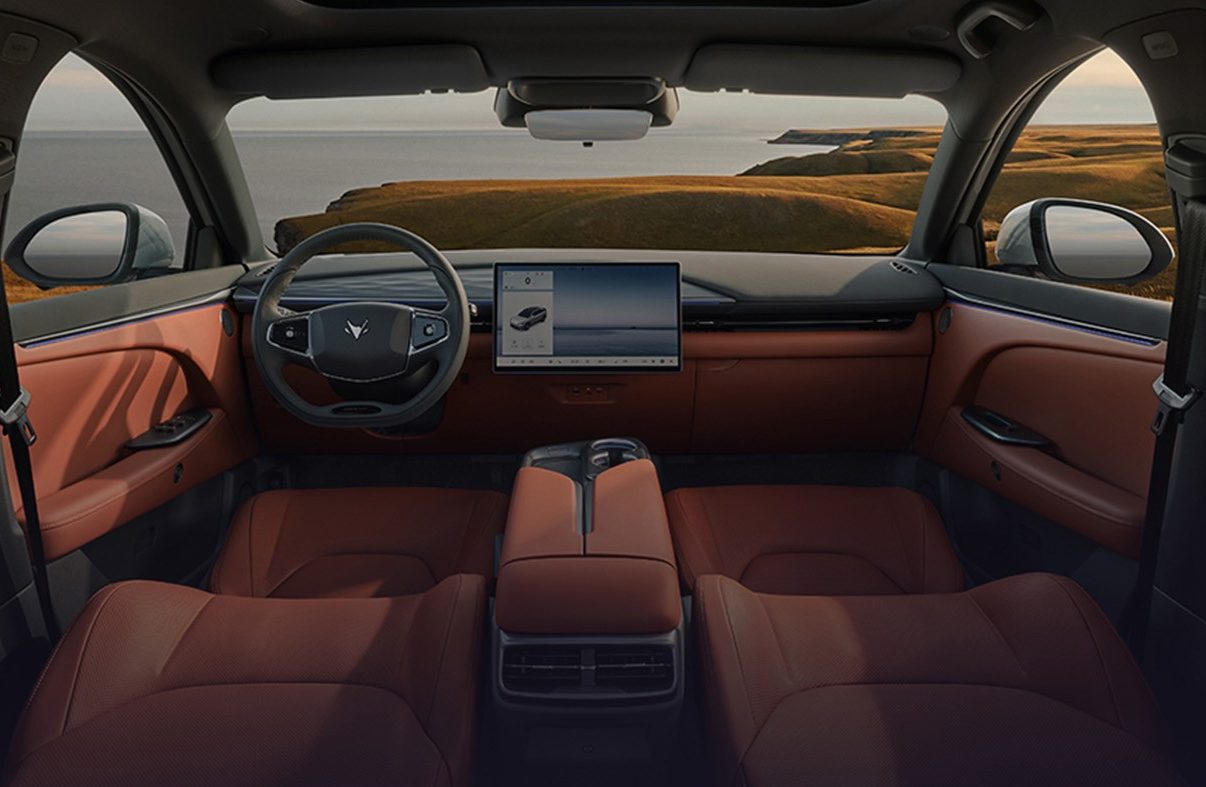 Interior de carro moderno com bancos e pain&eacute;is de porta em couro marrom, teto solar panor&acirc;mico e tela multim&iacute;dia. Ao fundo, paisagem costeira com mar e colinas gramadas sob c&eacute;u azul com nuvens esparsas
