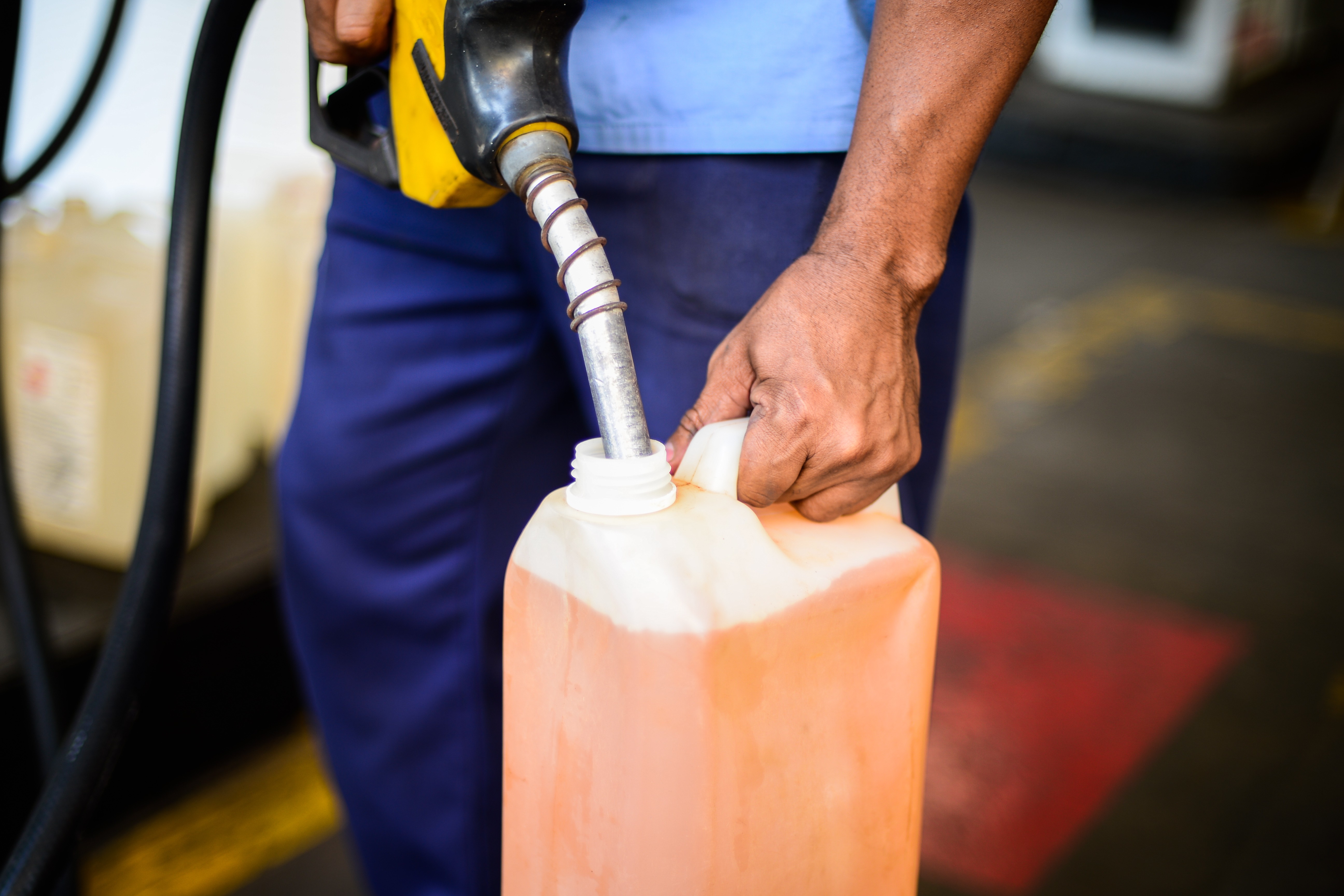Frentista com uniforme azul, segurando um bico de bomba de combust&iacute;vel amarelo e preto, abastecendo um gal&atilde;o pl&aacute;stico laranja com l&iacute;quido da mesma cor