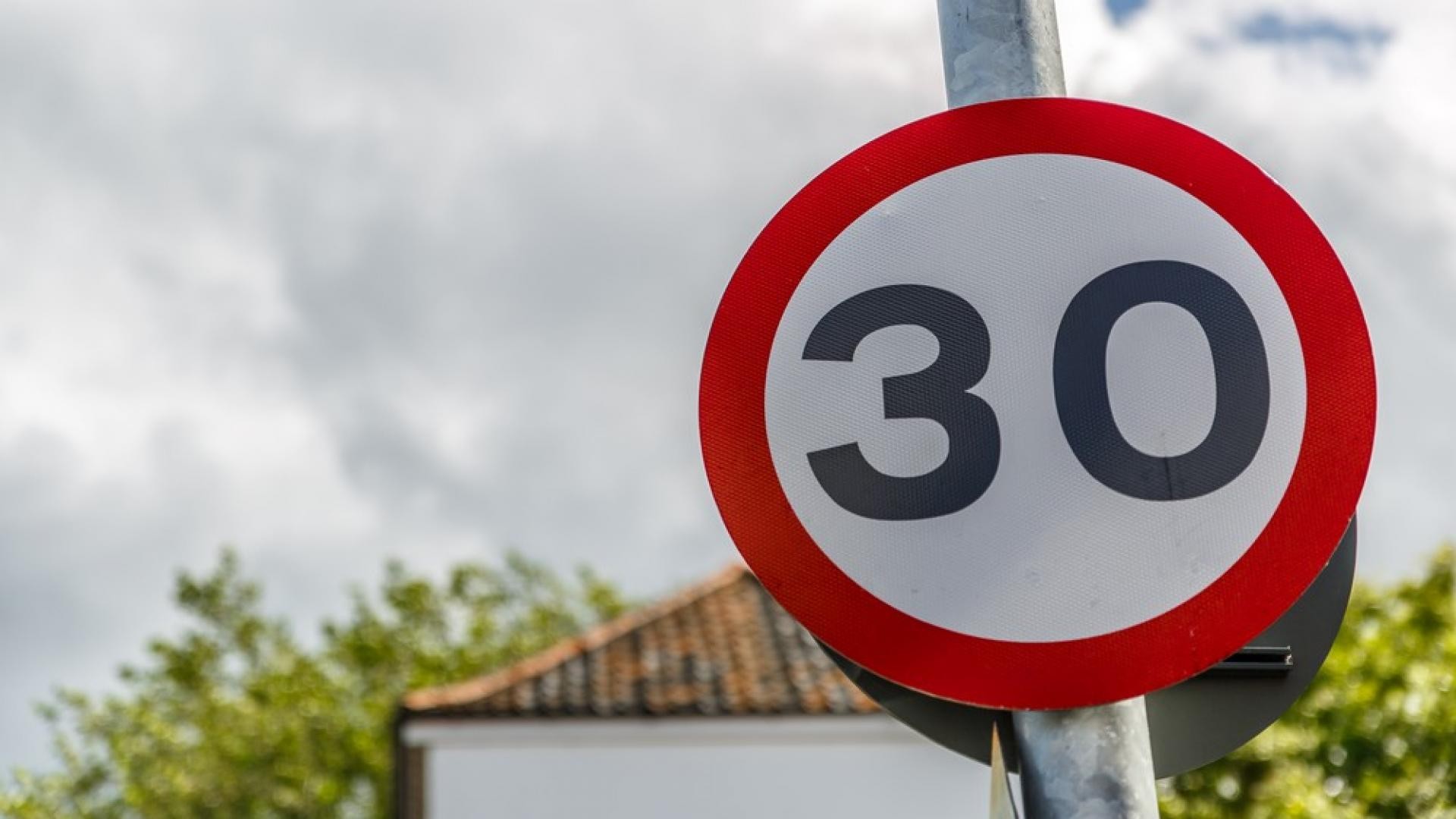 Placa de trânsito redonda com borda vermelha e fundo branco, exibindo o número 30 em preto, indicando limite de velocidade. Ao fundo, céu nublado e telhado de uma casa com árvores verdes