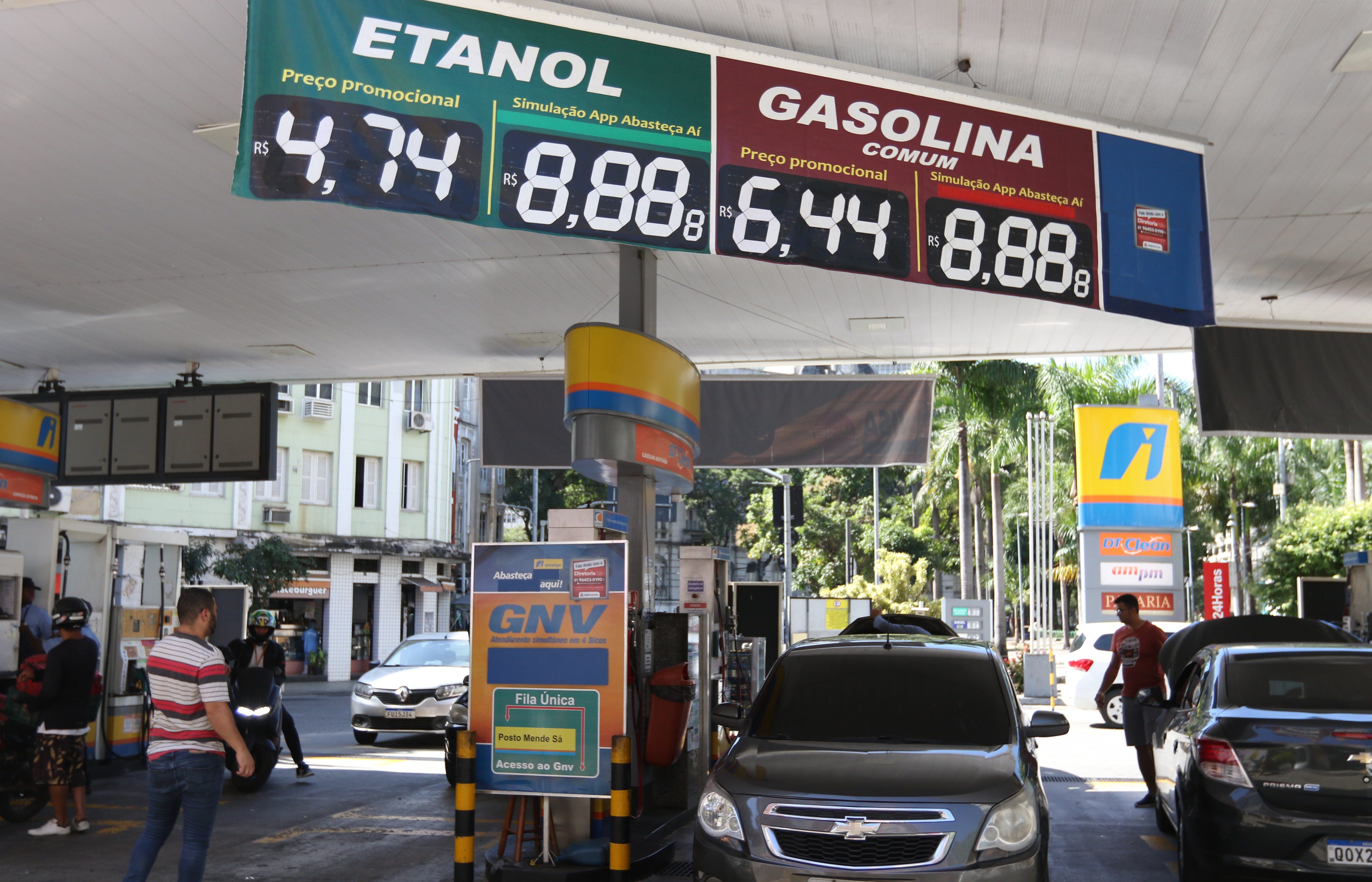 Posto de gasolina com letreiros digitais exibindo pre&ccedil;os: Etanol a R$ 4,74 e Gasolina Comum a R$ 6,44. Carros e pessoas circulam no local, com um painel de GNV &agrave; direita e pr&eacute;dios ao fundo