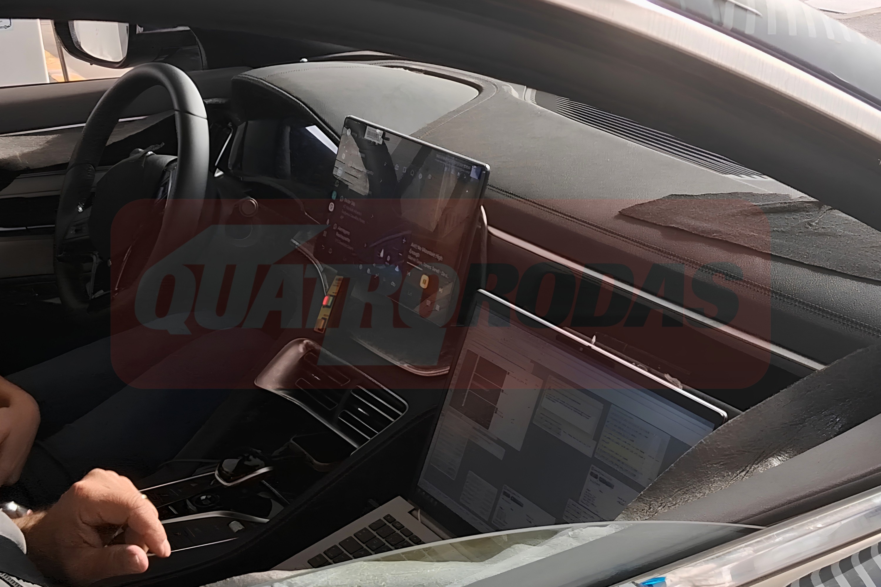 Interior de um carro com volante &agrave; esquerda, painel escuro e um tablet vertical no centro. Um notebook prateado est&aacute; aberto no banco do passageiro, refletindo a luz. Uma m&atilde;o masculina aparece no canto inferior esquerdo. A marca d&aacute;gua QUATRO RODAS est&aacute; sobreposta