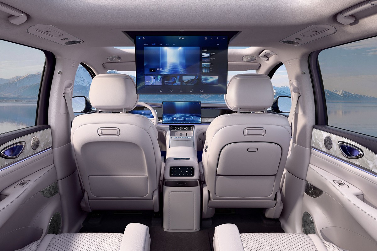 Interior de um carro de luxo com bancos claros, console central e duas telas digitais grandes. Uma tela retr&aacute;til no teto exibe um menu de entretenimento, e outra no painel mostra uma paisagem futurista. Ao fundo, montanhas e um lago azul.