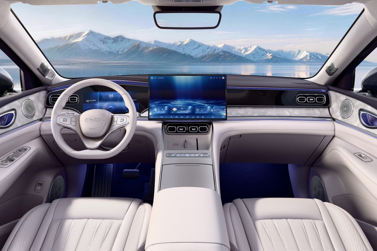 Interior de carro moderno com bancos de couro branco, volante claro e painel com tela grande exibindo uma imagem abstrata azul. Ao fundo, montanhas nevadas e um lago azul sob c&eacute;u claro, vistos pelo para-brisa