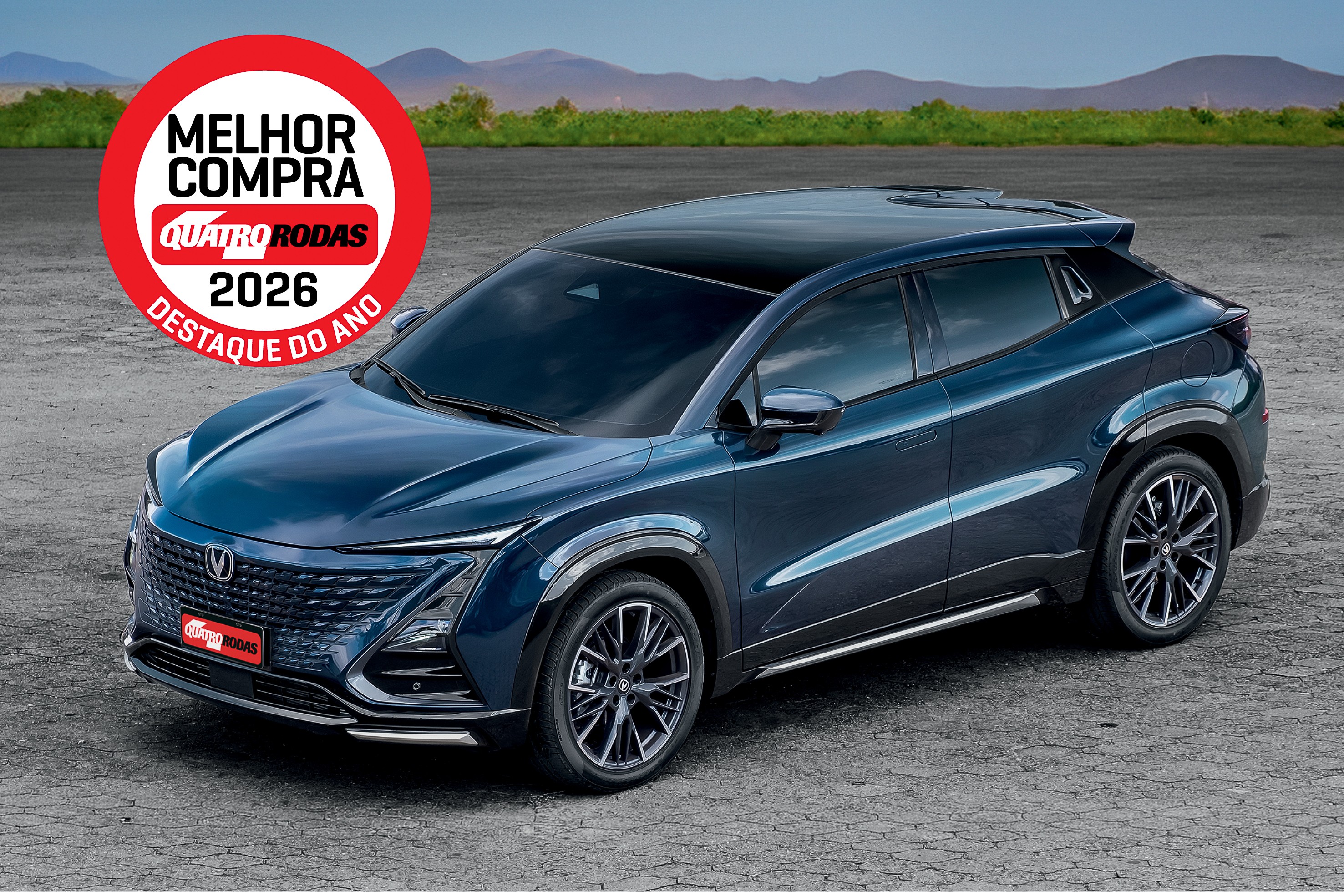 SUV azul escuro, com teto preto e rodas esportivas, estacionado em uma superf&iacute;cie cinza. No canto superior esquerdo, um selo vermelho e branco indica MELHOR COMPRA QUATRO RODAS 2026 DESTAQUE DO ANO. Ao fundo, montanhas e vegeta&ccedil;&atilde;o verde sob um c&eacute;u azul
