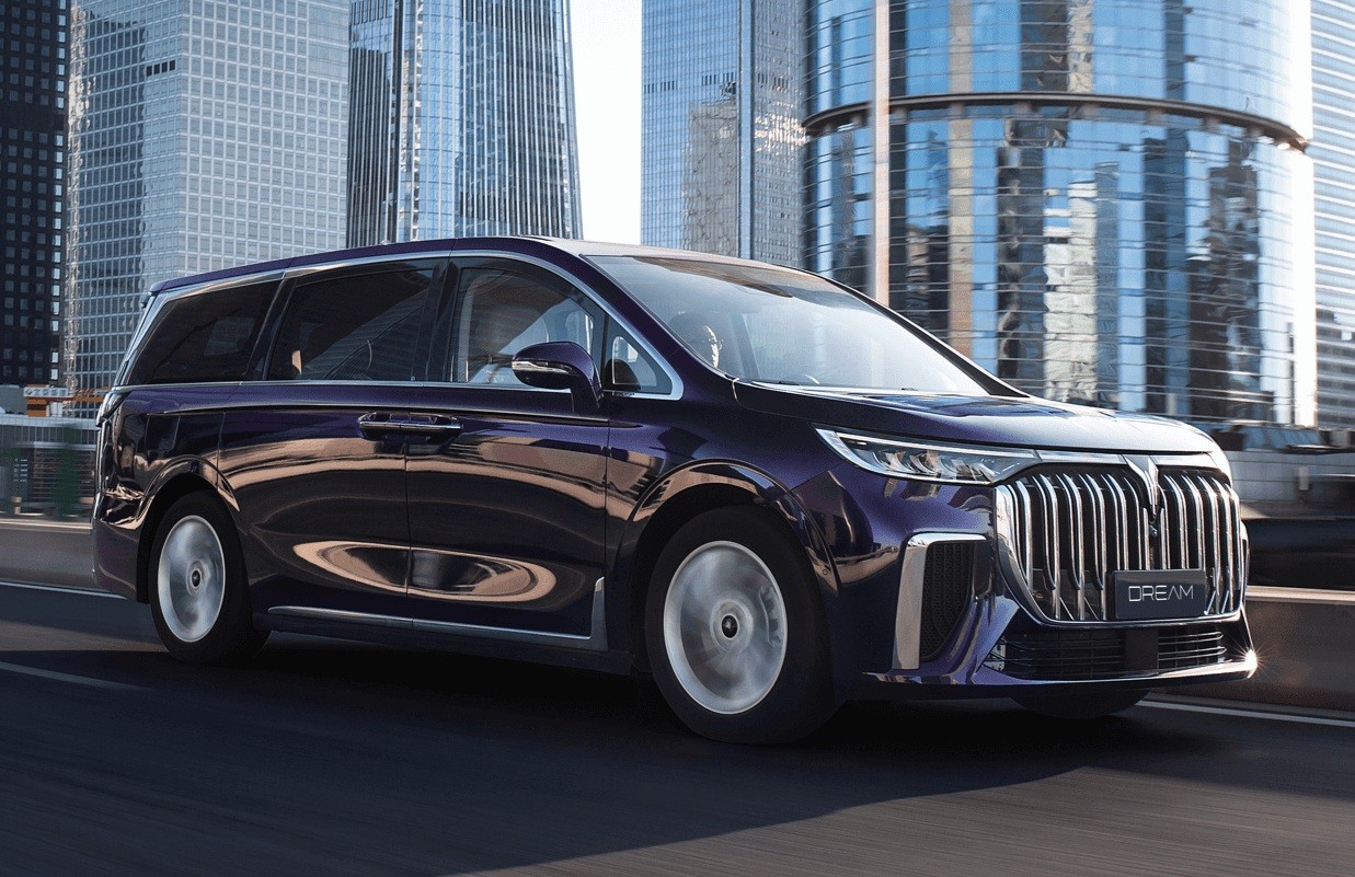 Minivan roxa escura, modelo Dream, em movimento numa via urbana, com pr&eacute;dios modernos de vidro ao fundo