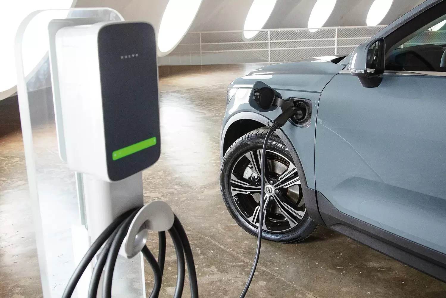 Carro elétrico cinza-azulado sendo carregado em estação de recarga branca com detalhes verdes, em ambiente interno com piso de concreto e teto curvo