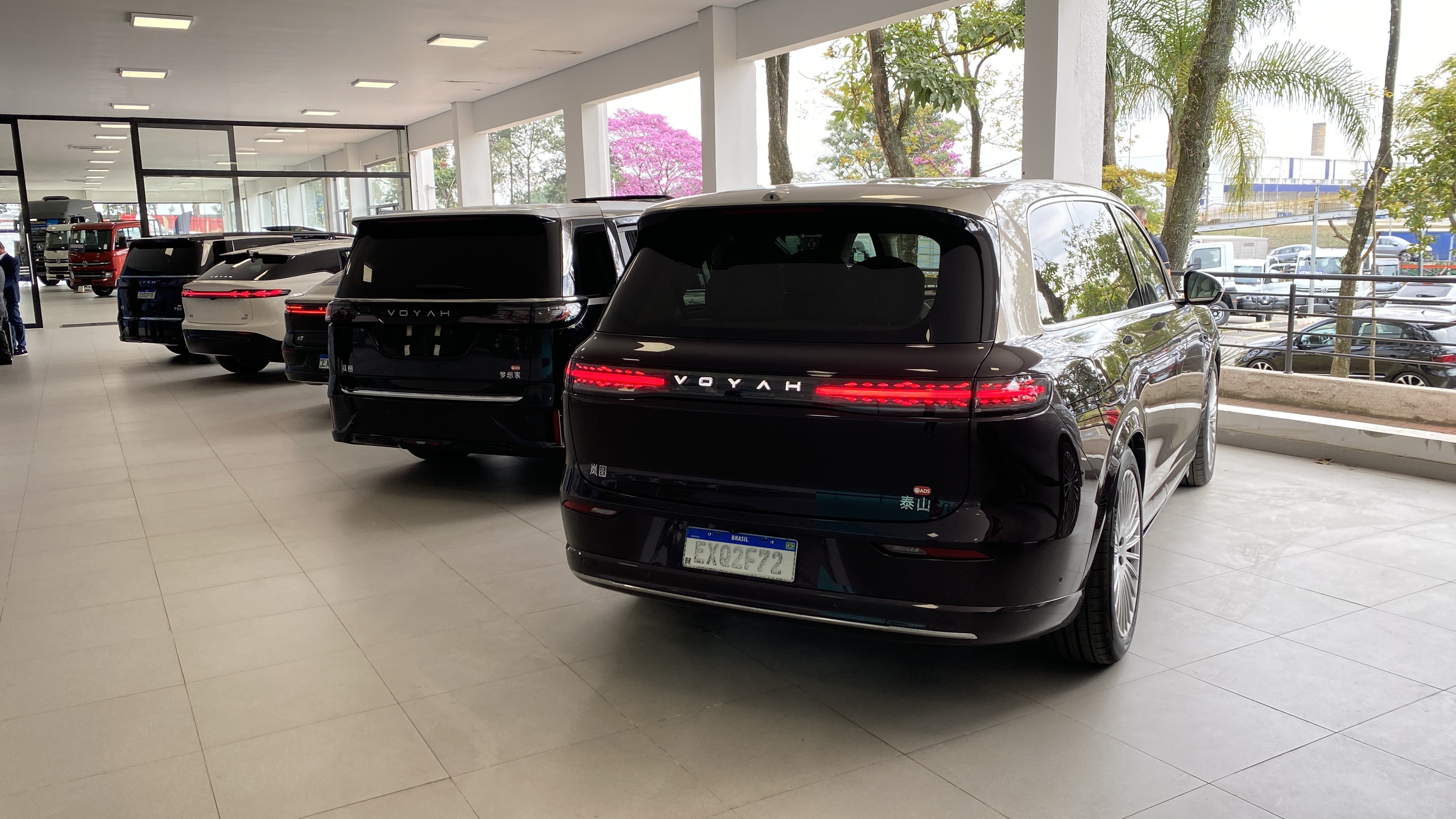 V&aacute;rios carros de luxo, incluindo um SUV preto Voyah com placa brasileira, estacionados em uma concession&aacute;ria com janelas grandes e &aacute;rvores ao fundo
